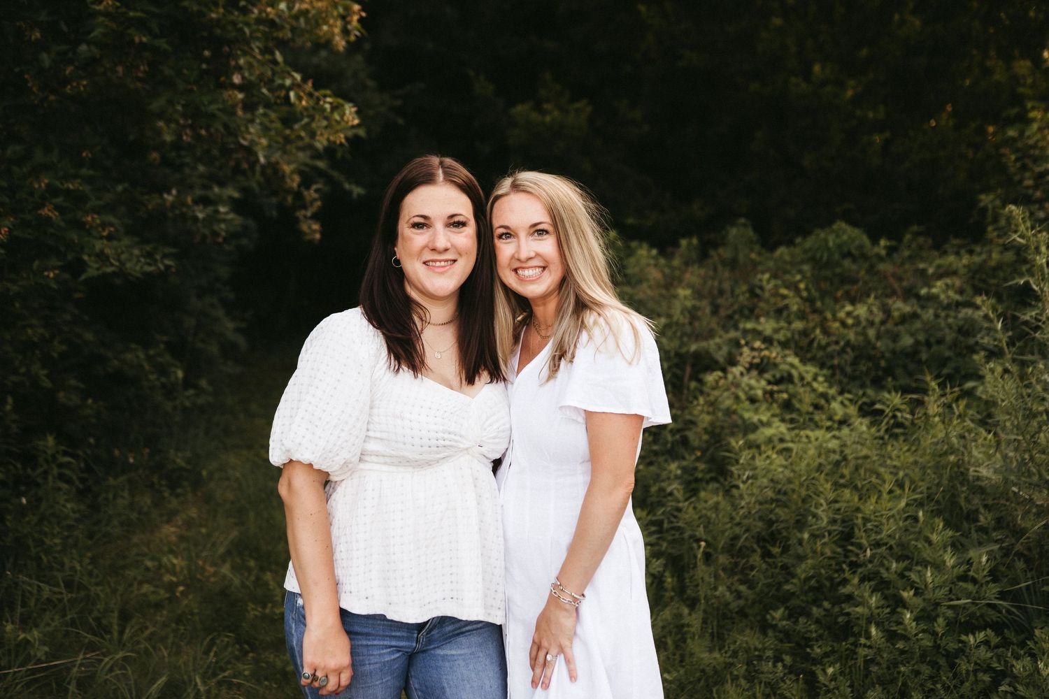 Two women wearing white tops stand together outdoors with greenery in the background.