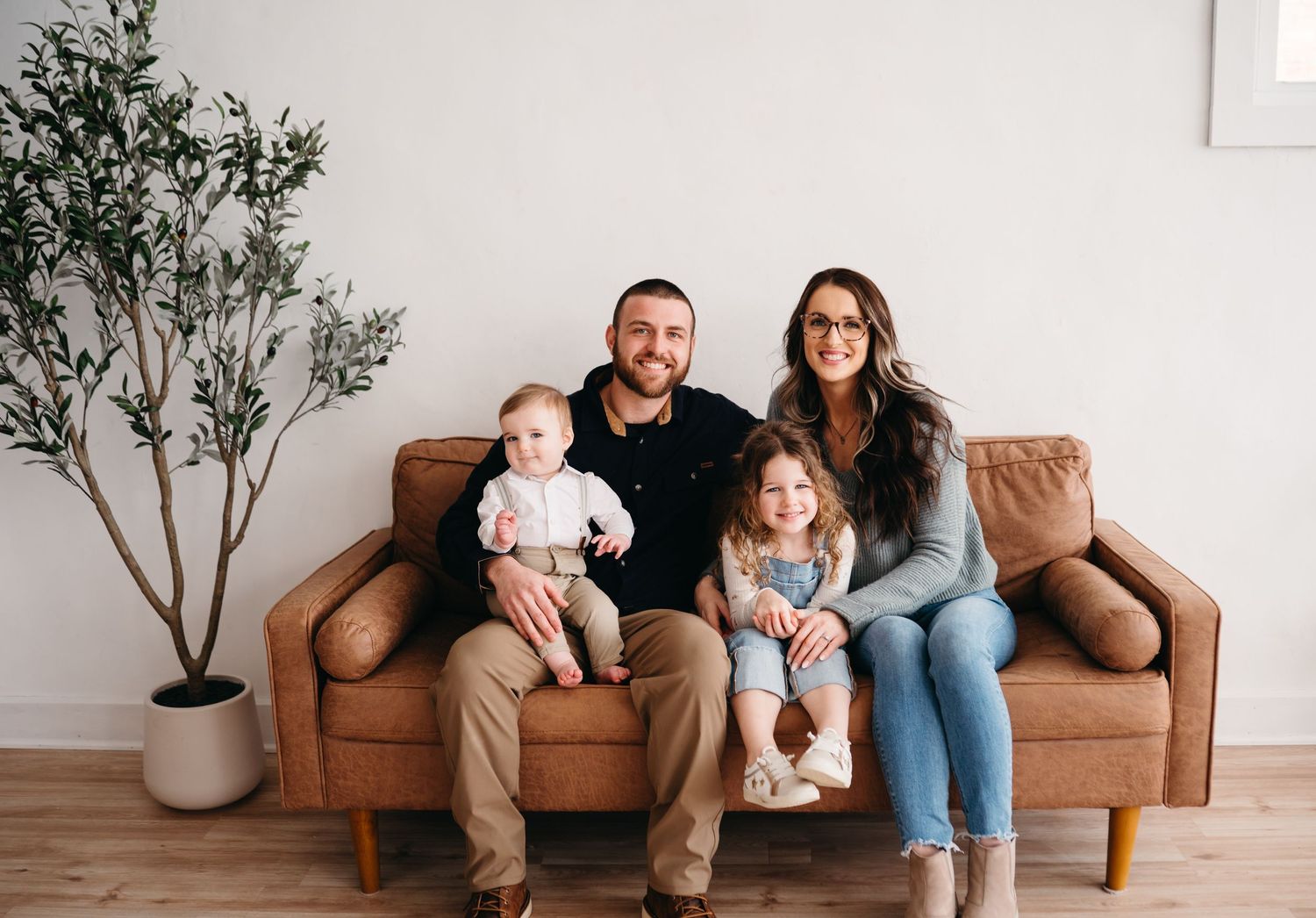 Casual family portrait on tan leather sofa with decorative plant in modern setting.