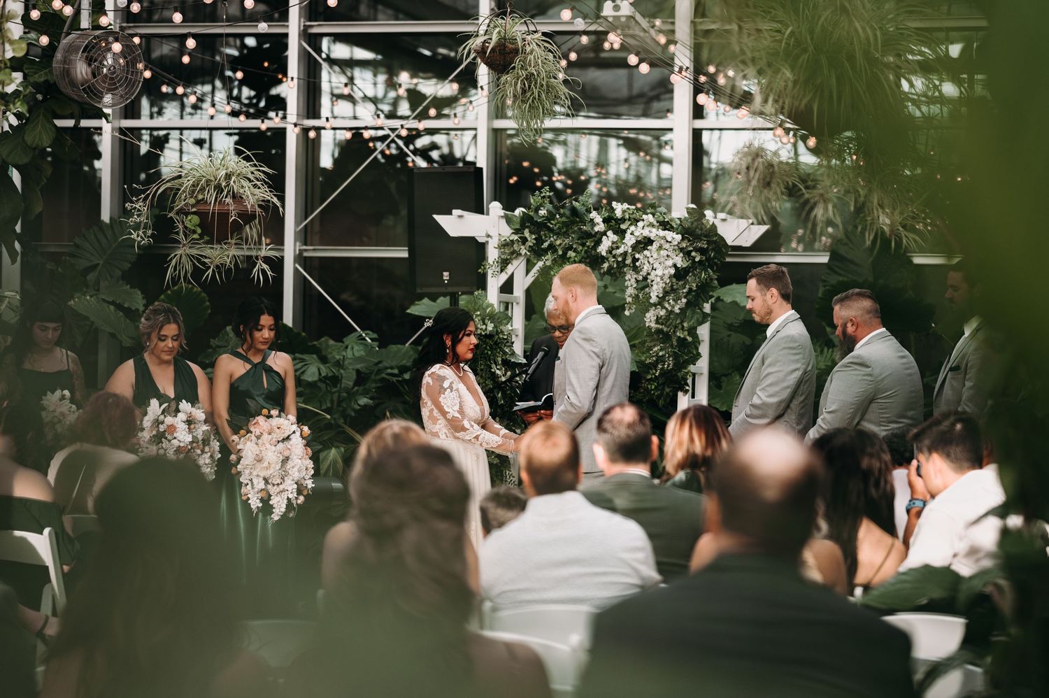 Wedding ceremony in a greenhouse venue with string lights and tropical plants creating an intimate atmosphere.
