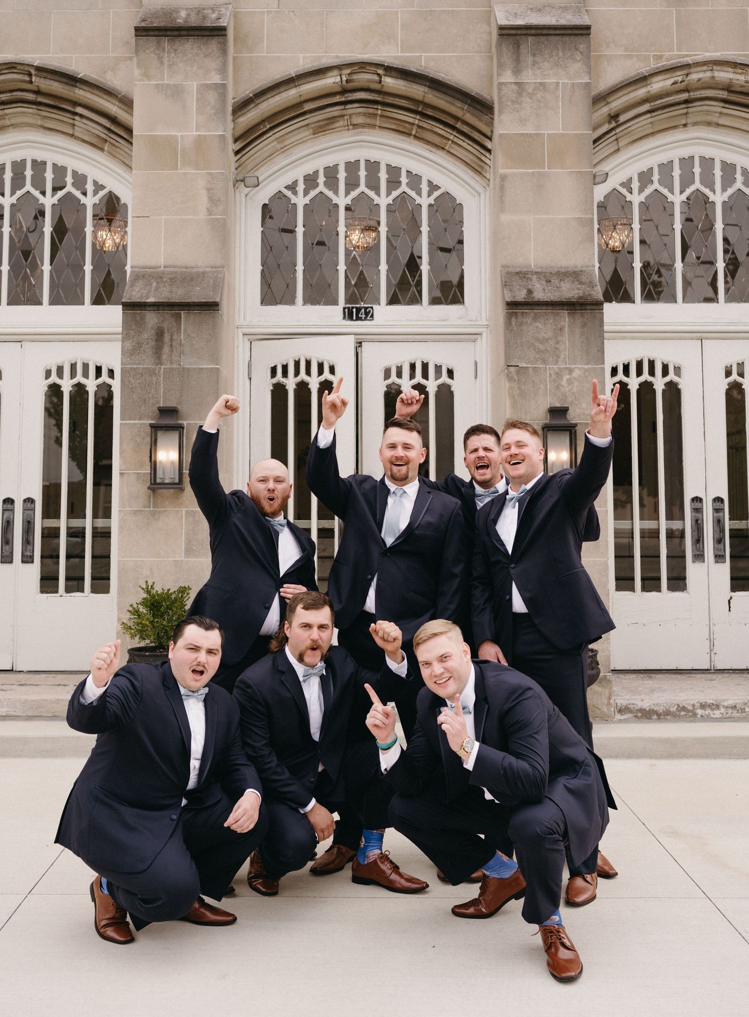 Groomsmen group showing excitement with energetic poses outside church entrance.
