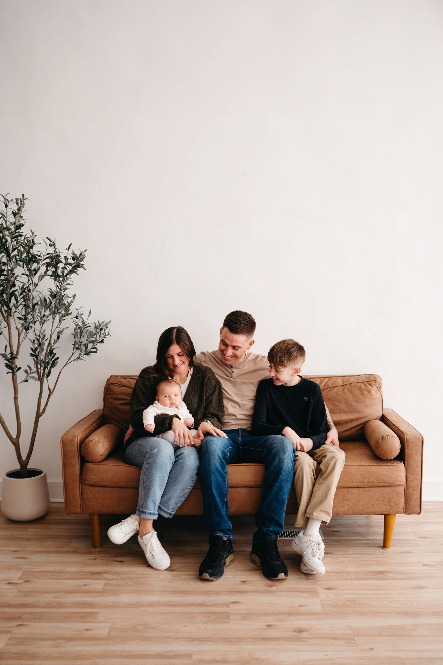 A family sits together on a brown leather couch in a minimalist room with a potted plant.