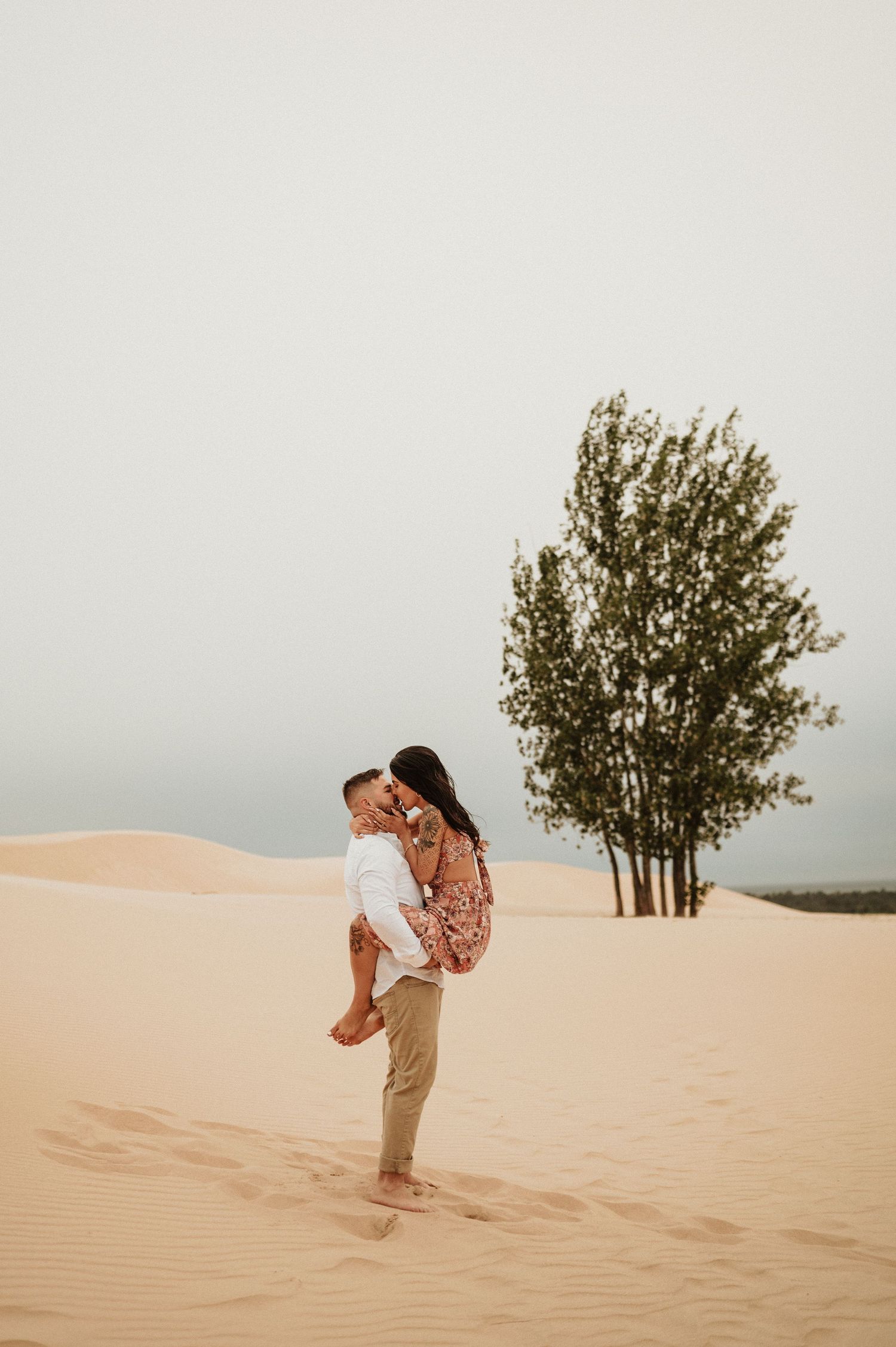 A romantic couple embraces on sandy dunes with trees in the background during golden hour.