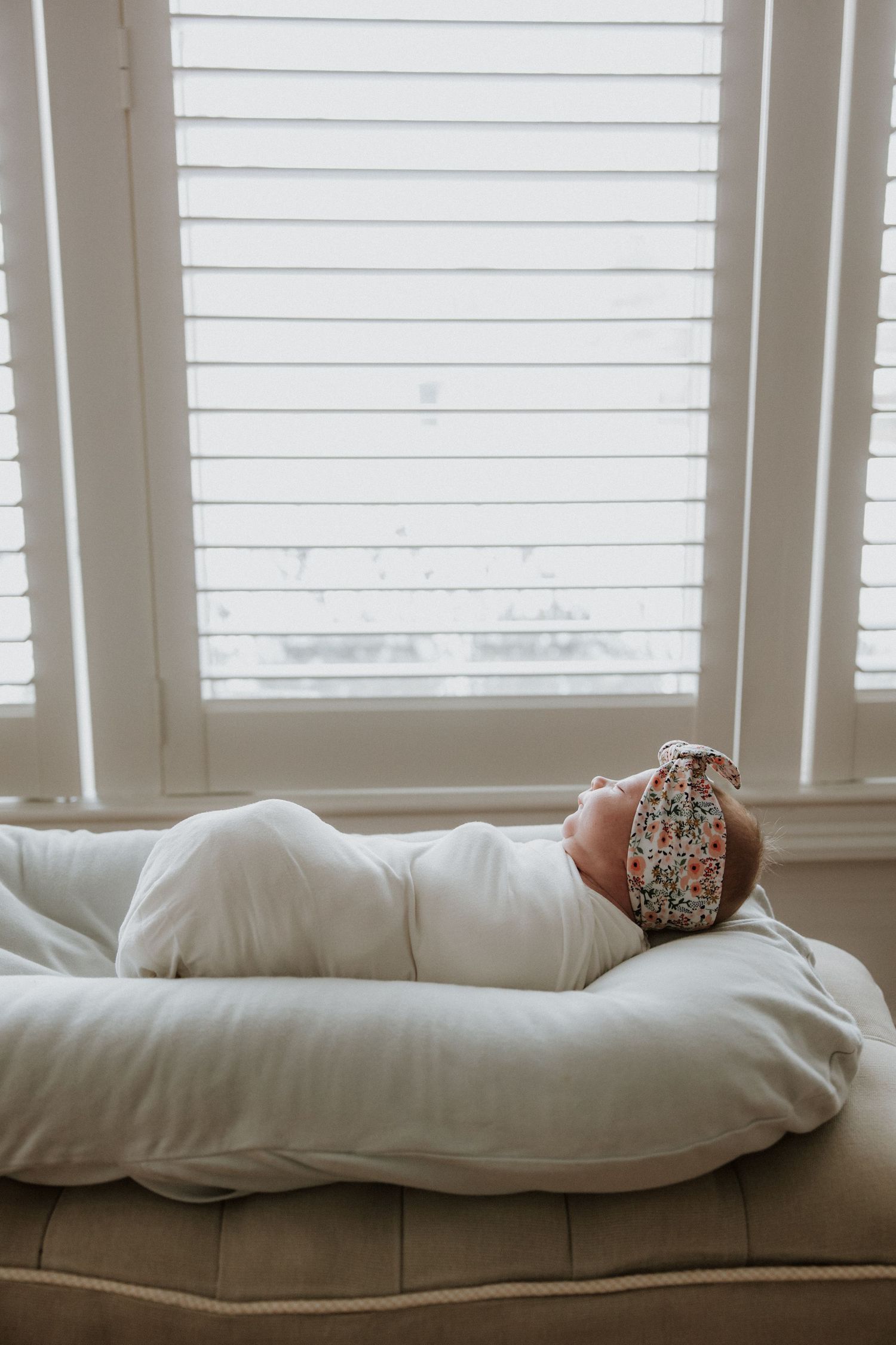 Newborn baby in white swaddle rests peacefully on nursing pillow by bright window.