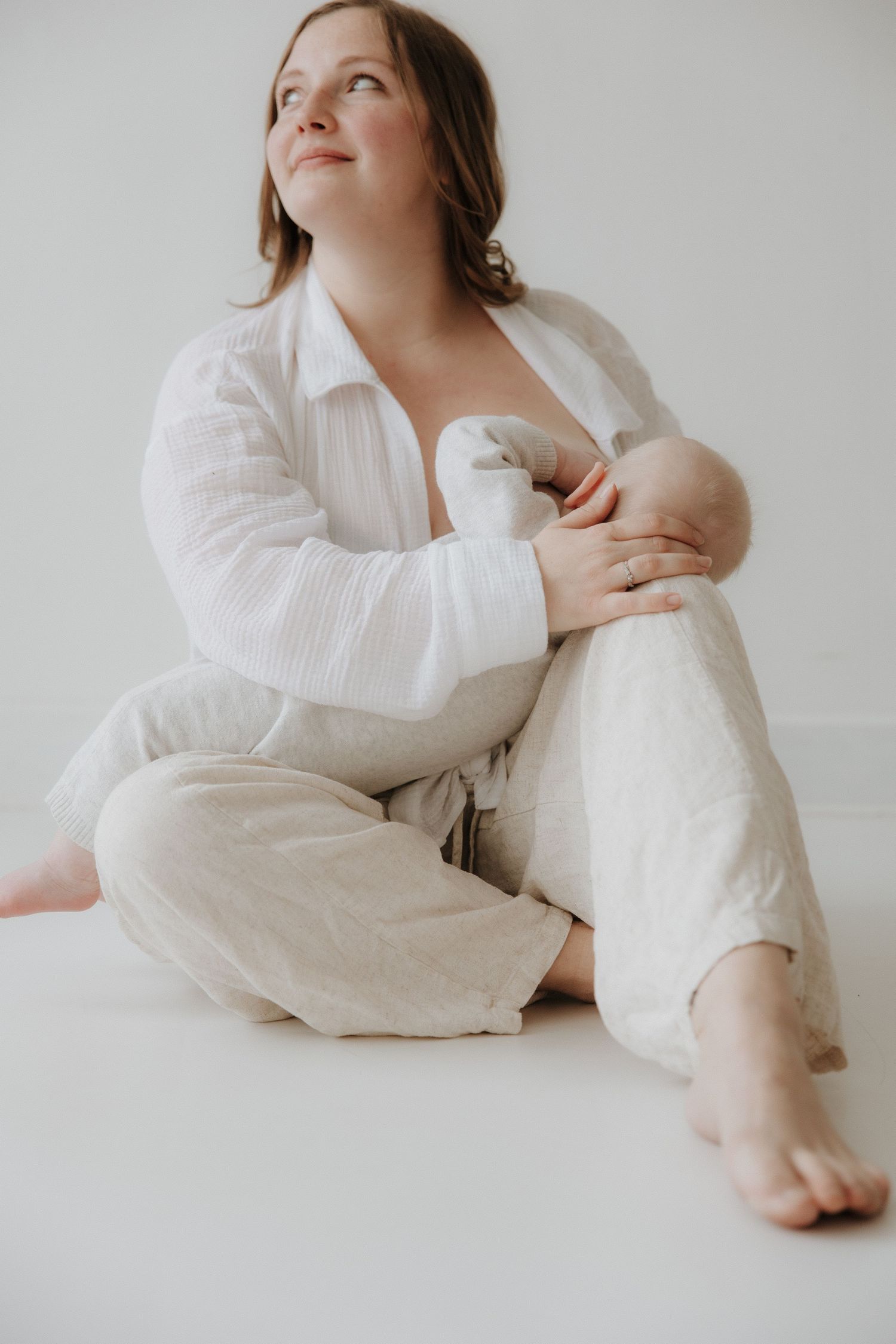 Barefoot mother in white shirt and linen pants breastfeeding newborn while sitting.