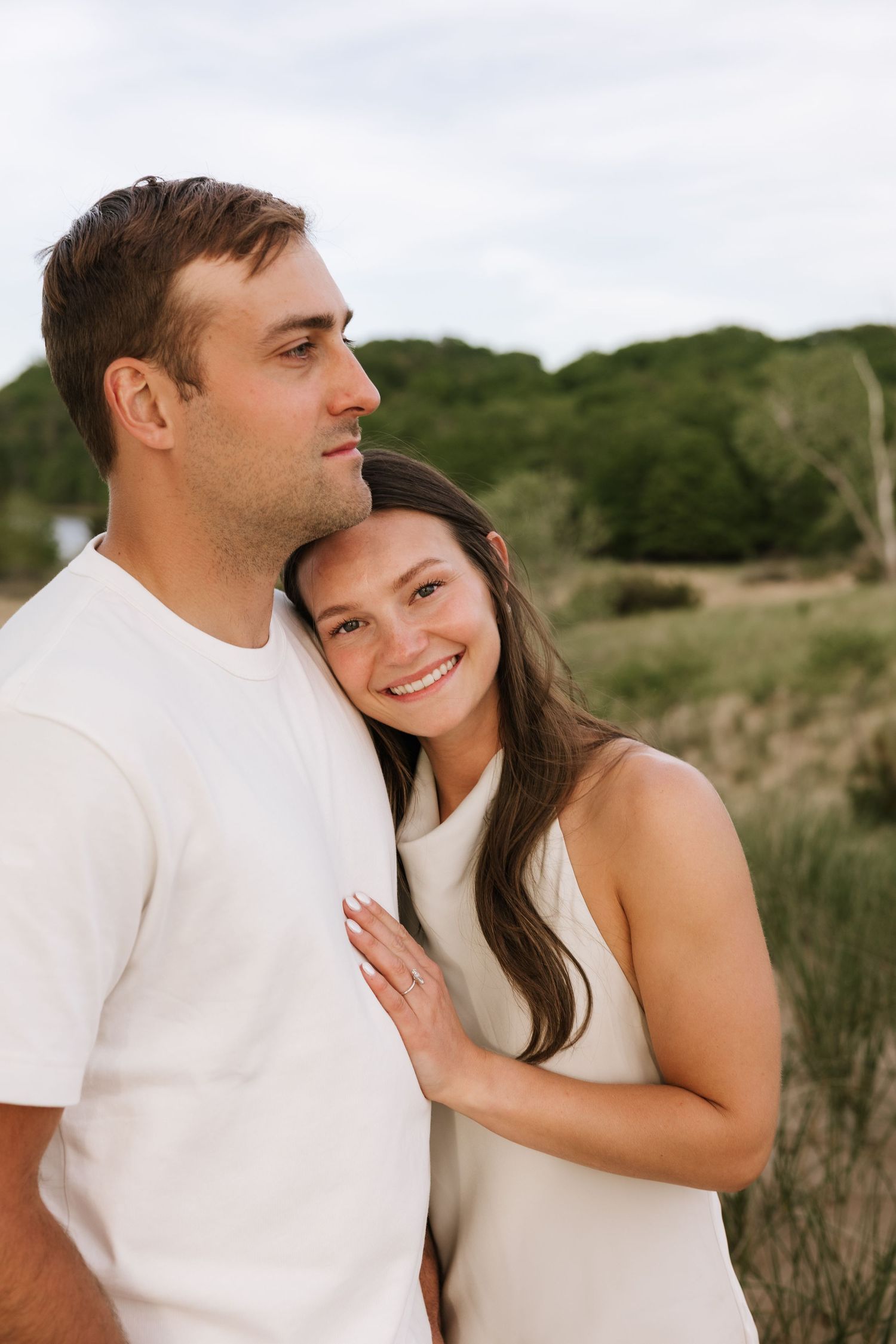 Couple cuddling outdoors in grassy field, woman smiling at camera while leaning against mans shoulder.