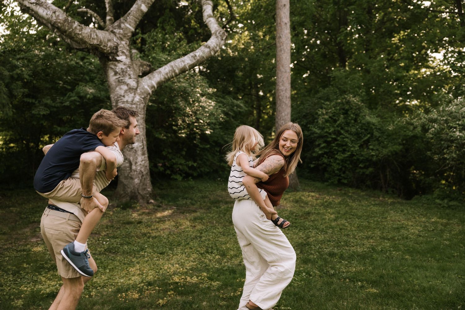 Family playing outdoors in lush green yard, parents swinging laughing children among trees on sunny day.