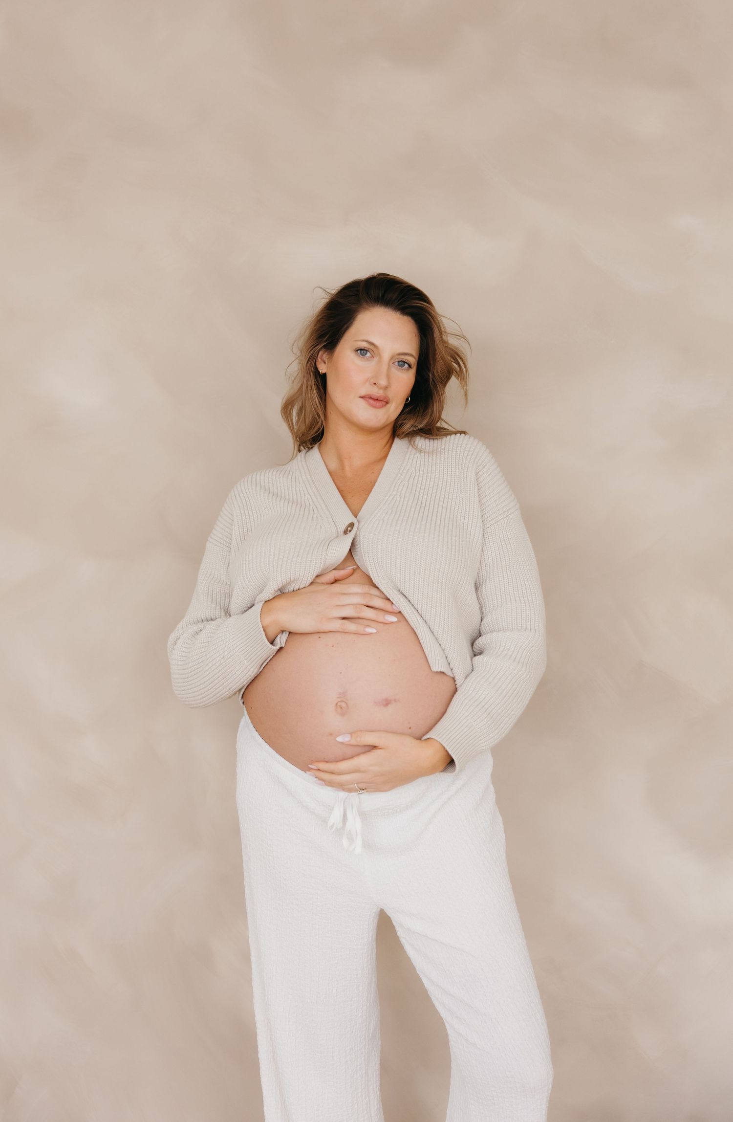 Pregnant woman in open beige knit cardigan and white pants cradles her bare bump against a warm beige backdrop.