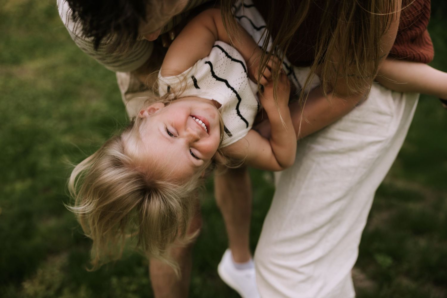 A joyful child laughs upside down while being held by a parent outdoors on green grass.