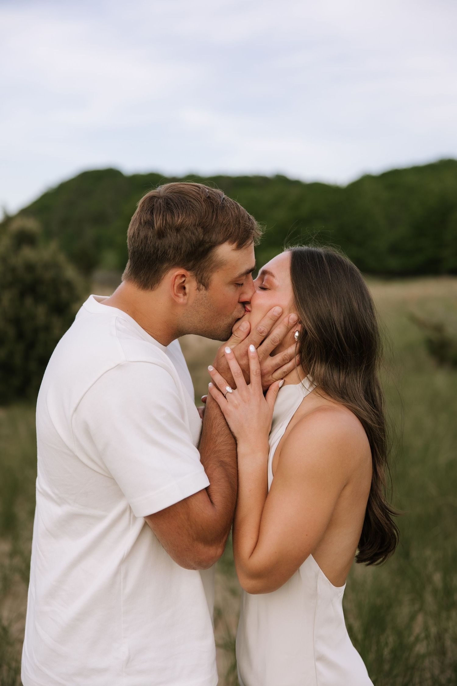 Couple sharing a romantic kiss in a green field, man tenderly holding woman's face, both wearing white outfits.