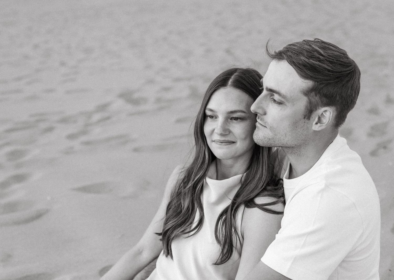 Black and white close-up of a young couple sitting together on sand, the man kissing the womans cheek.