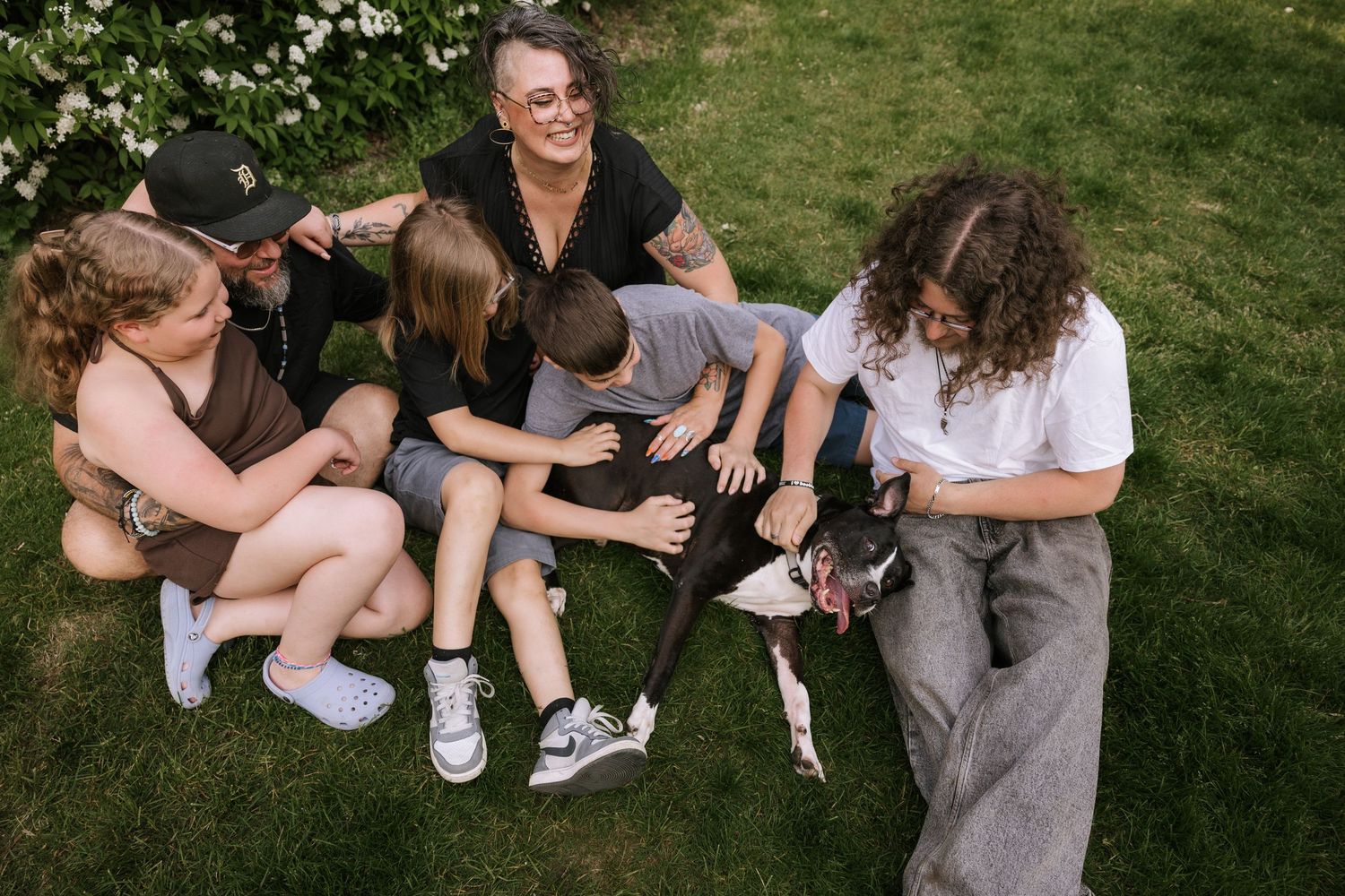 Happy family of five sitting on grass together petting their black and white pit bull dog.