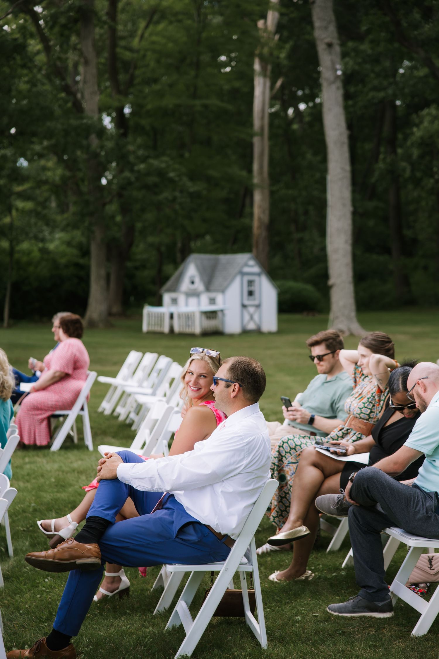 Wedding guests relaxing in white folding chairs on a lawn during outdoor ceremony.