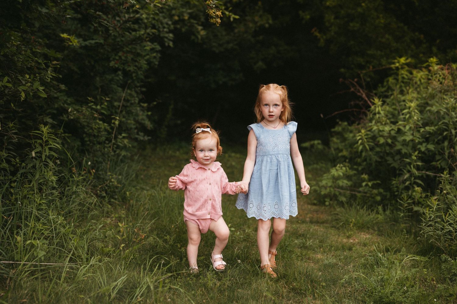 Two children walk hand in hand through a grass path surrounded by green foliage.