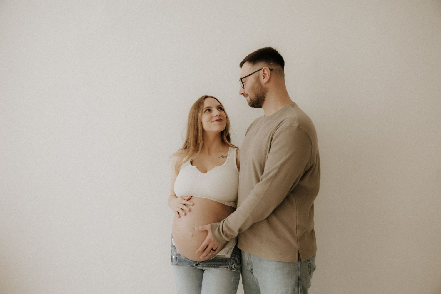 Couple in casual attire sharing an intimate moment against a neutral wall during a maternity photoshoot.