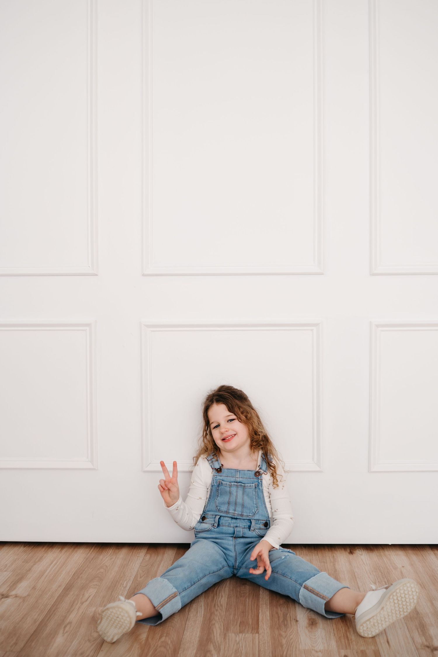 Child in denim overalls sits on hardwood floor against white wall making peace sign while smiling playfully.