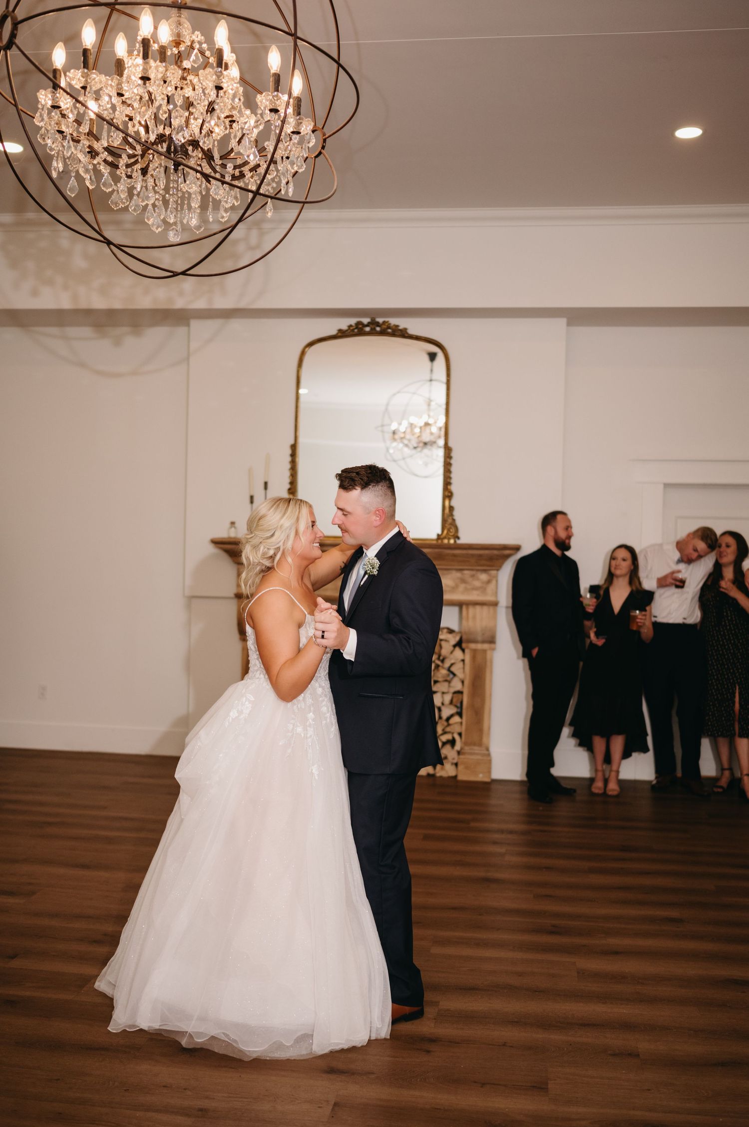 A wedding couple dances beneath a crystal chandelier in an elegant ballroom setting.
