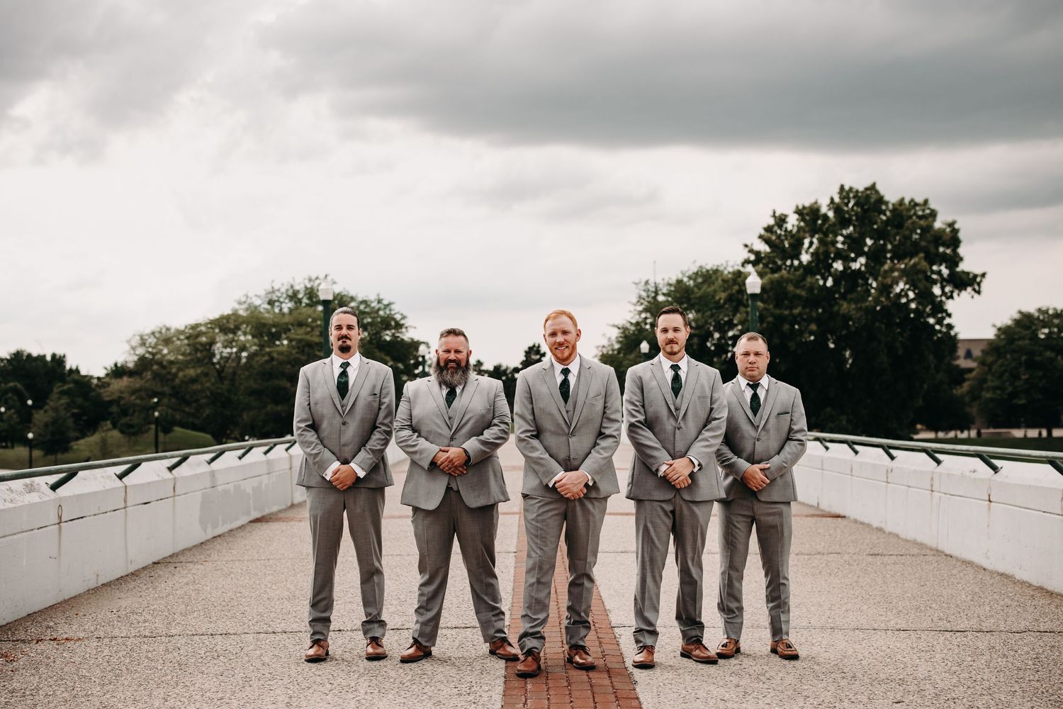 Groomsmen in matching gray suits pose on a white bridge under cloudy skies.
