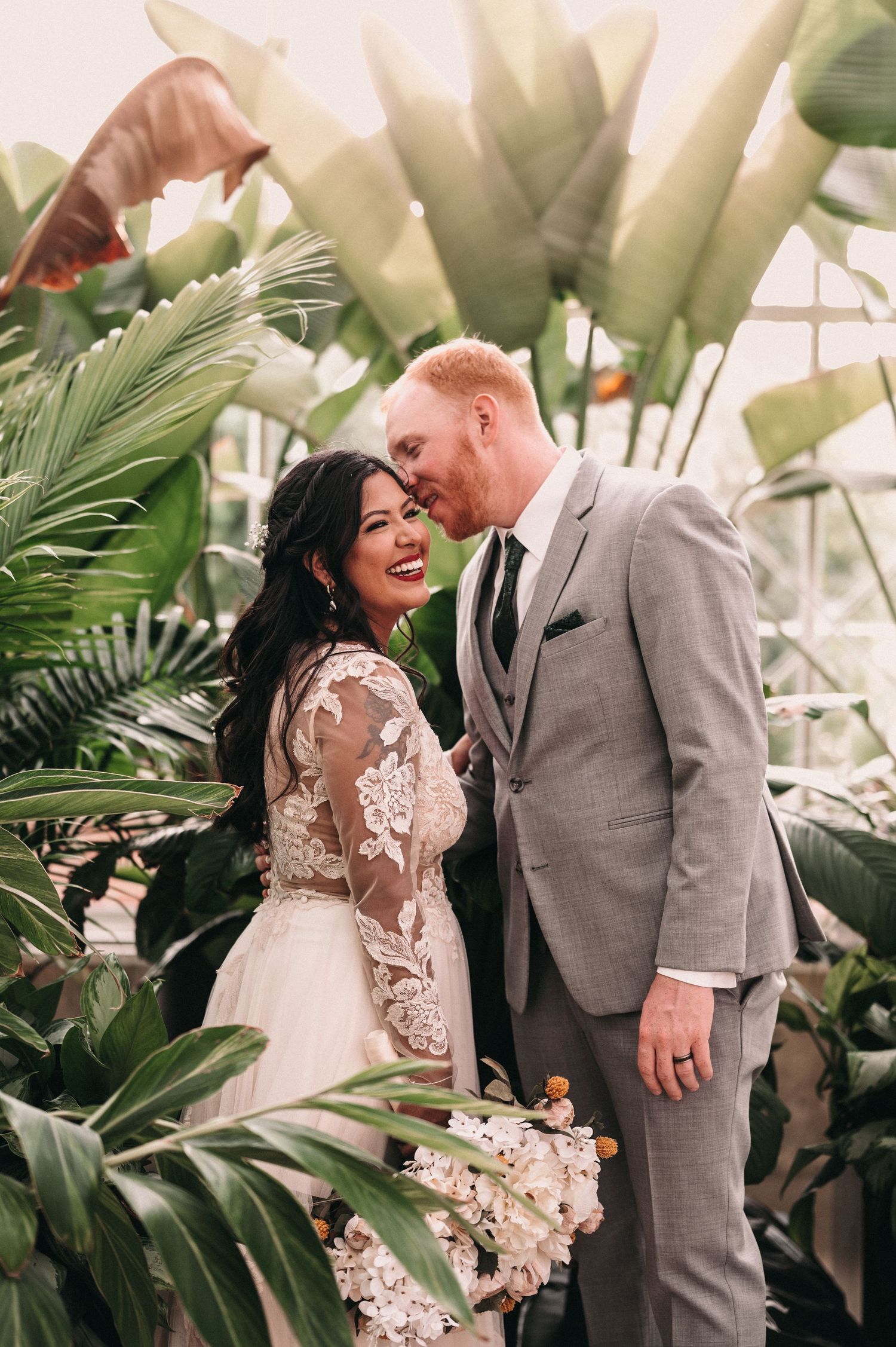 A romantic wedding portrait in a greenhouse with lush tropical plants and white flowers.