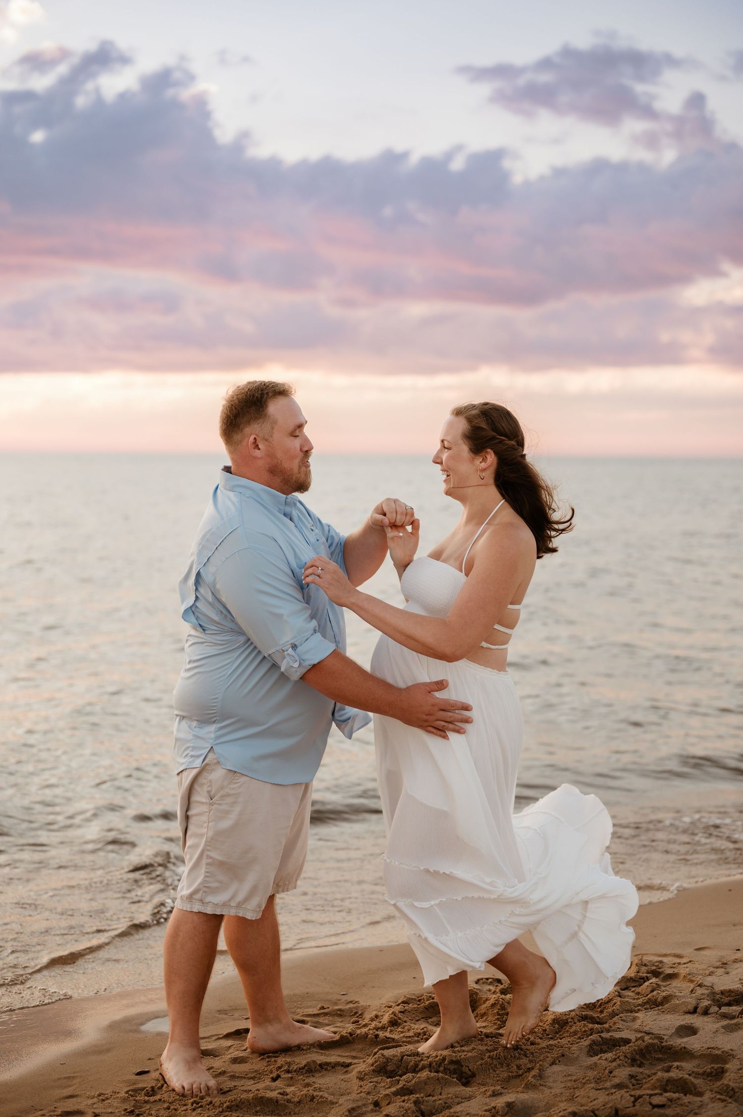 A couple dances together on a beach at sunset with pink clouds and ocean waves in the background.
