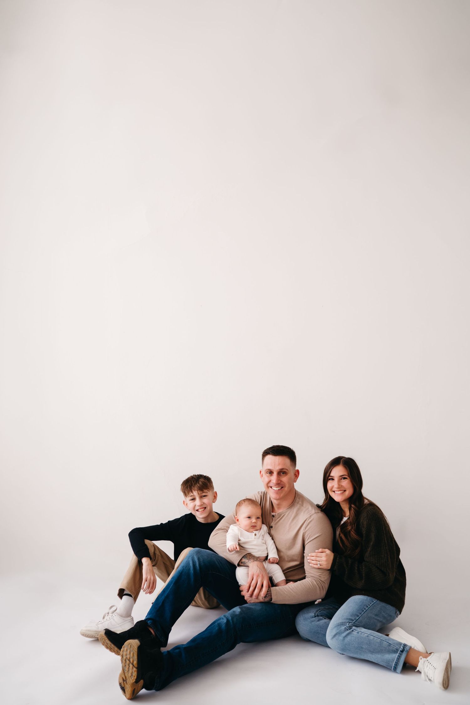 Family sits together on the floor in a minimal studio with white background in casual attire.