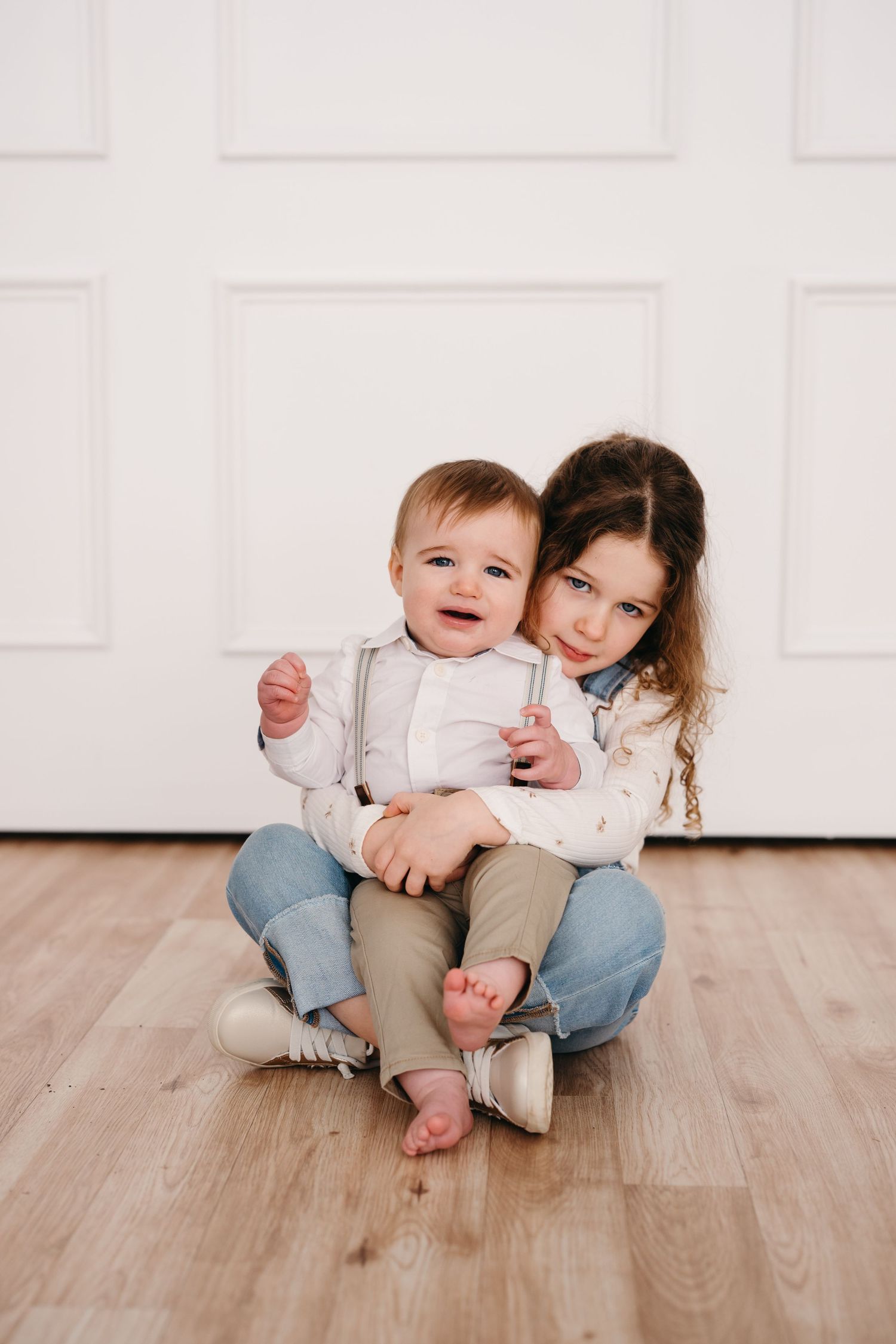 Two children sitting together on a hardwood floor against a white wall, sharing a sweet embrace.