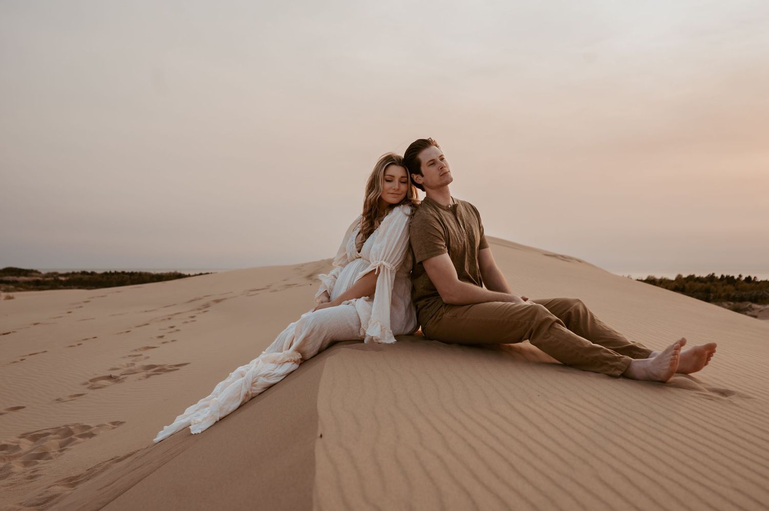 A couple sits together on a sand dune during sunset in desert landscape.