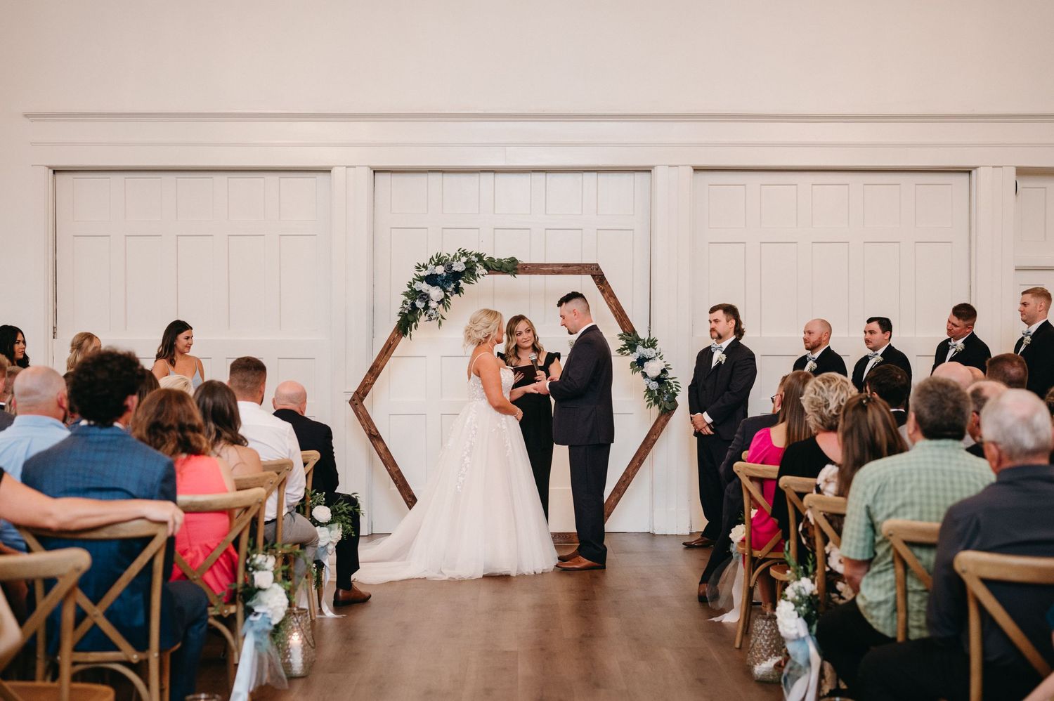Wedding ceremony in progress under a hexagonal arch with guests seated in cross-back chairs.