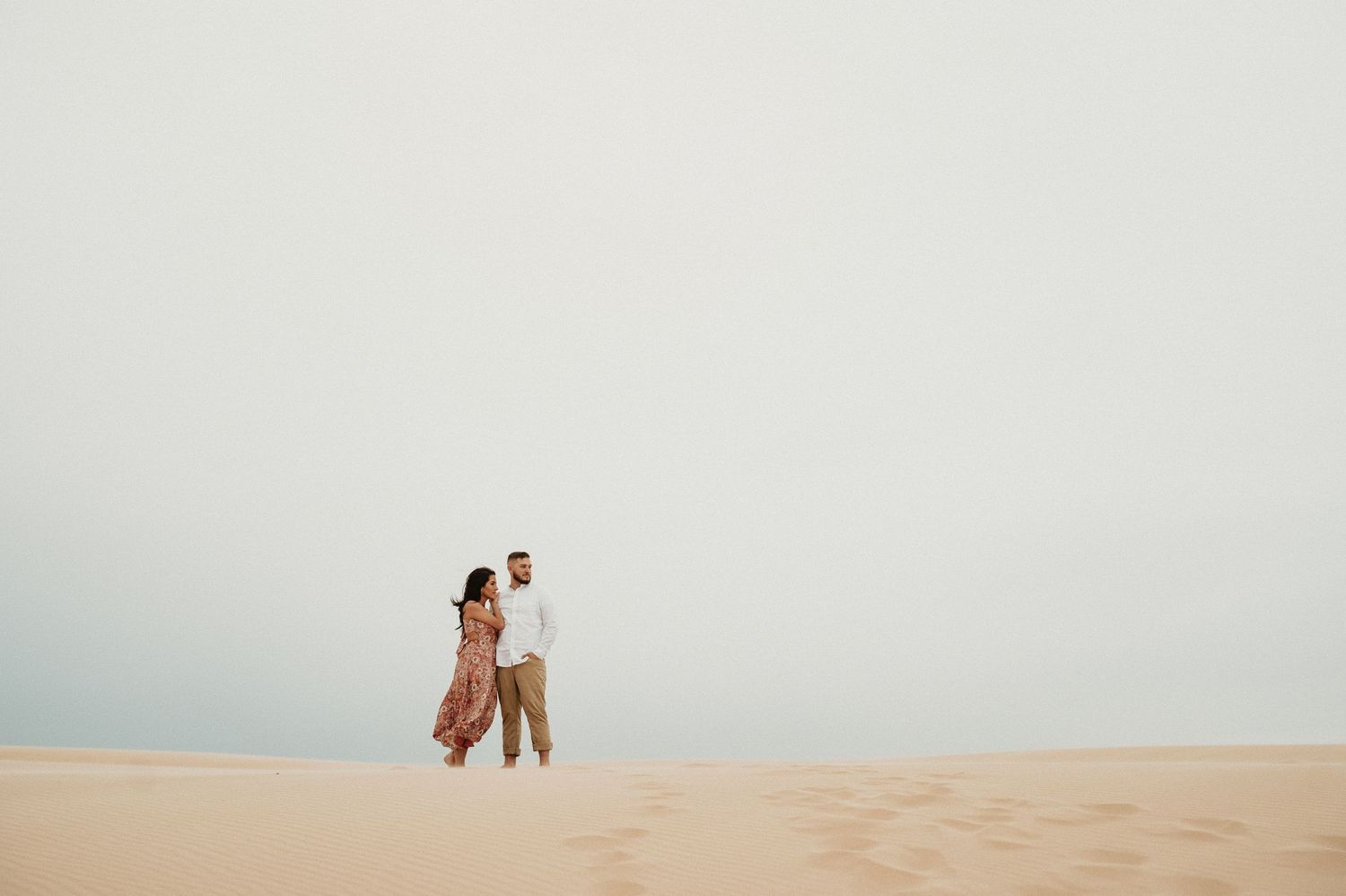 A couple walks together across vast desert sand dunes under cloudy skies.