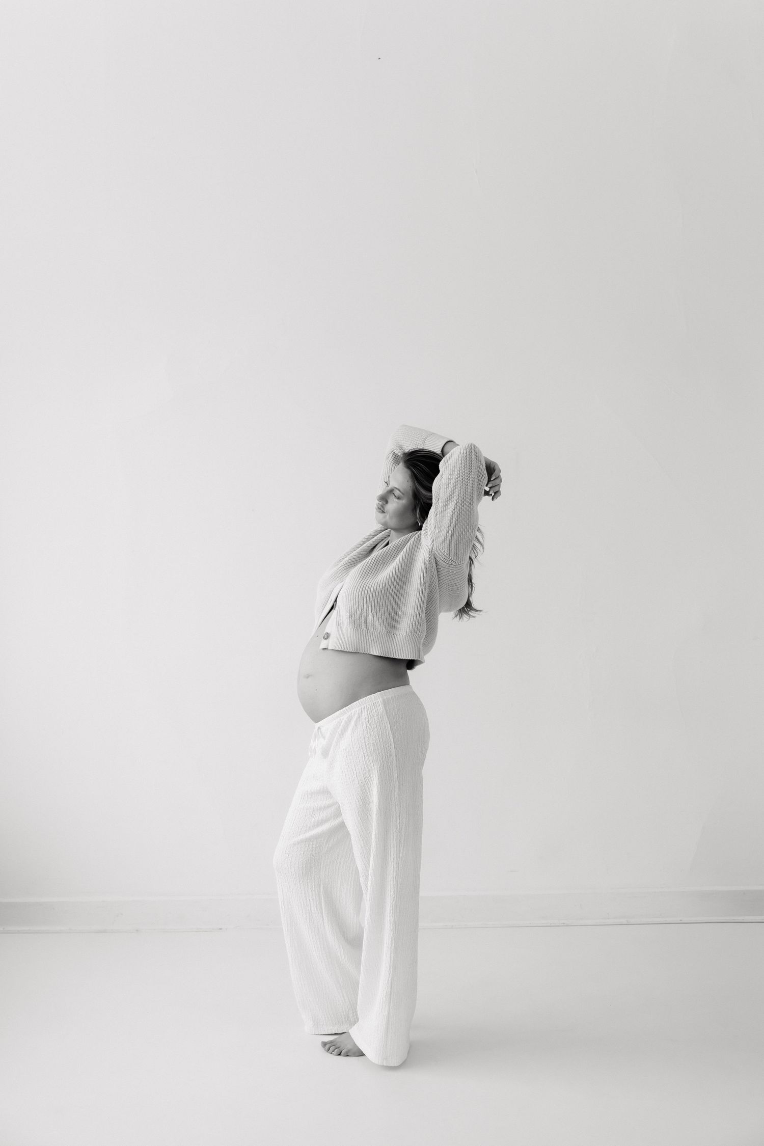 Black and white maternity photo of a pregnant woman standing sideways, arms raised above her head, showing her baby bump.