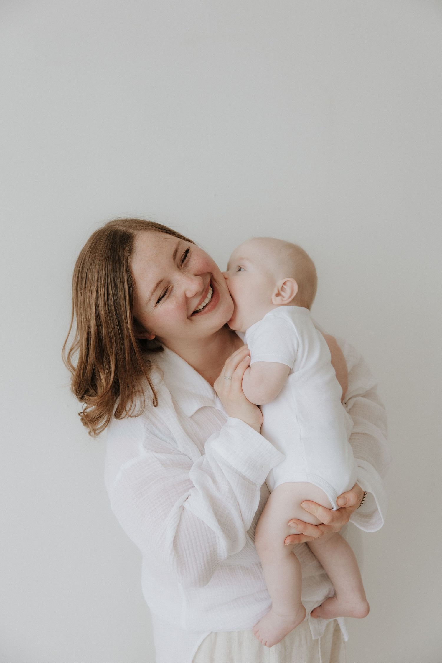 A joyful mother in a white shirt laughs while holding her smiling baby against a light background.