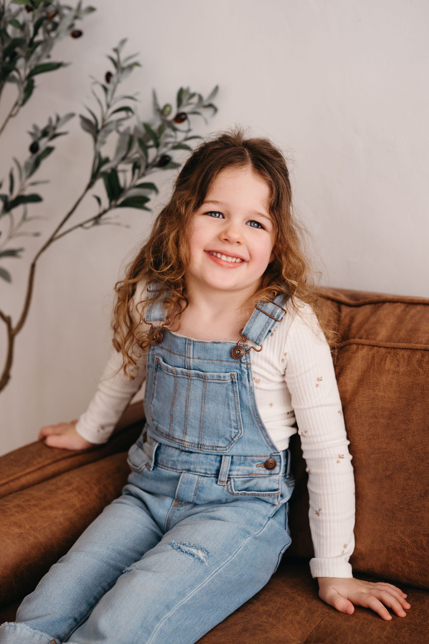 A young child wearing denim overalls and white sweater sits smiling on a brown leather couch near decorative foliage.