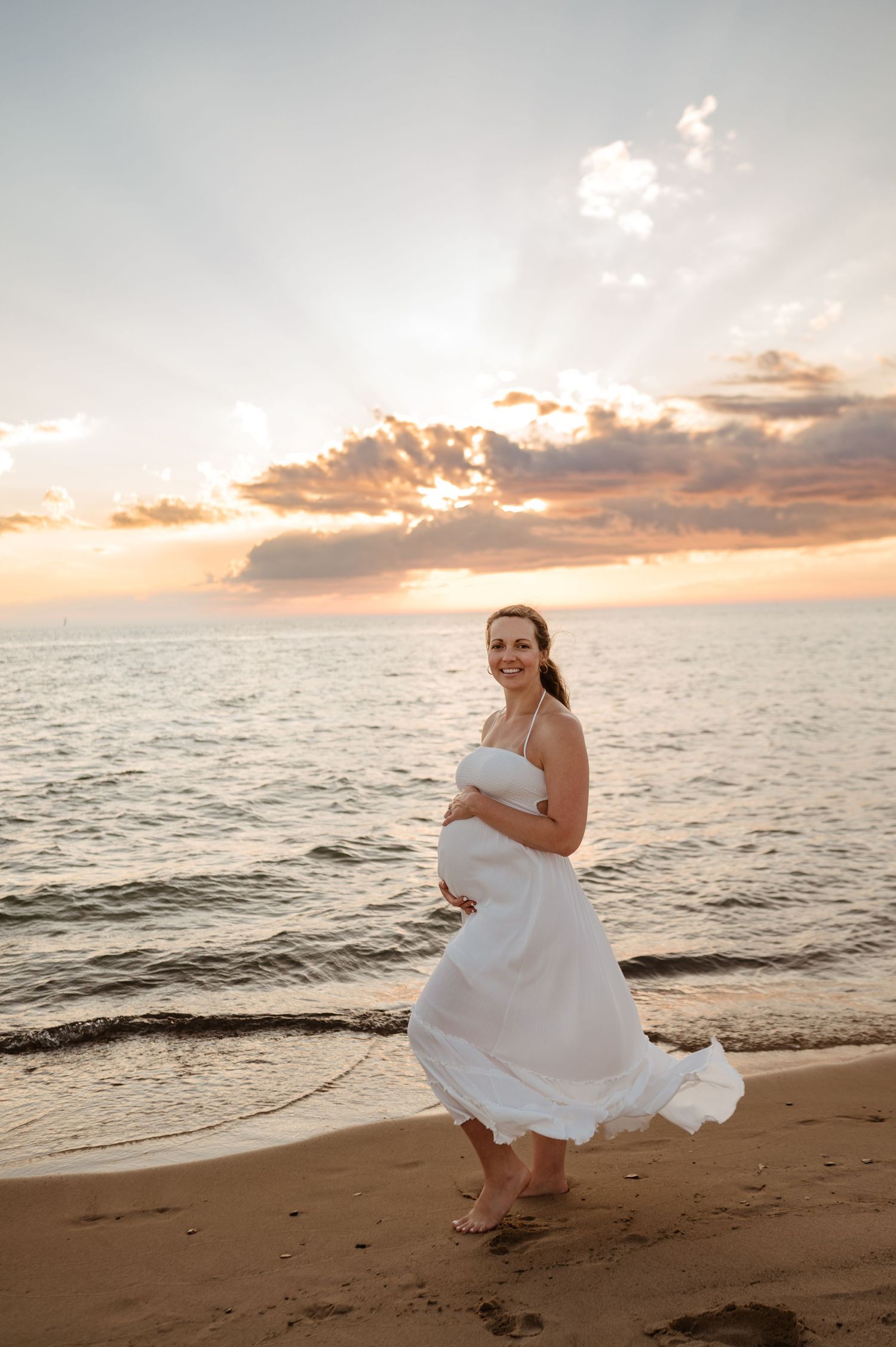 A person in a flowing white dress walks along a beach at sunset as golden light streams through clouds over the ocean.