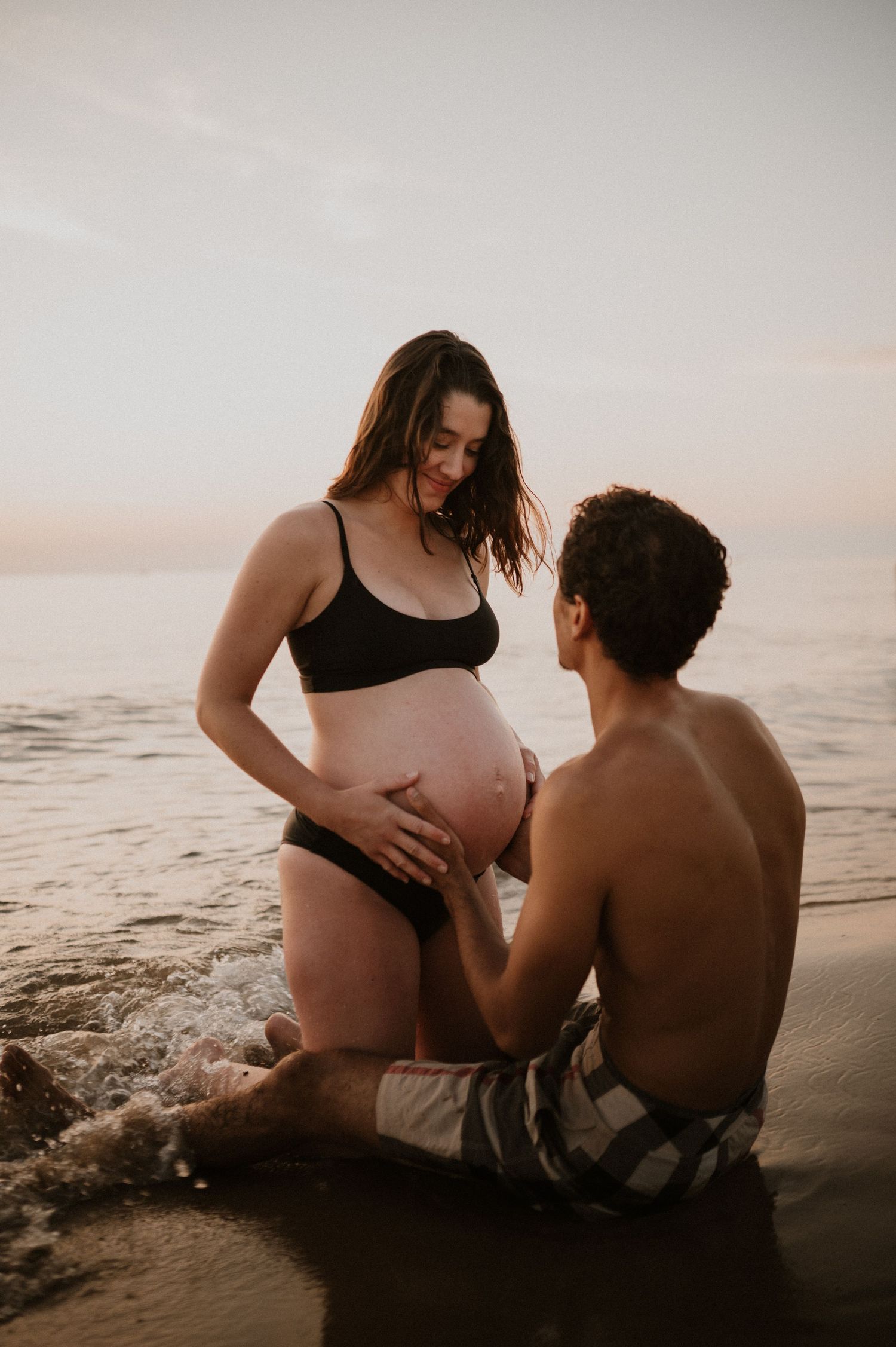 Intimate maternity photoshoot on beach at sunset with couple sitting in sand.