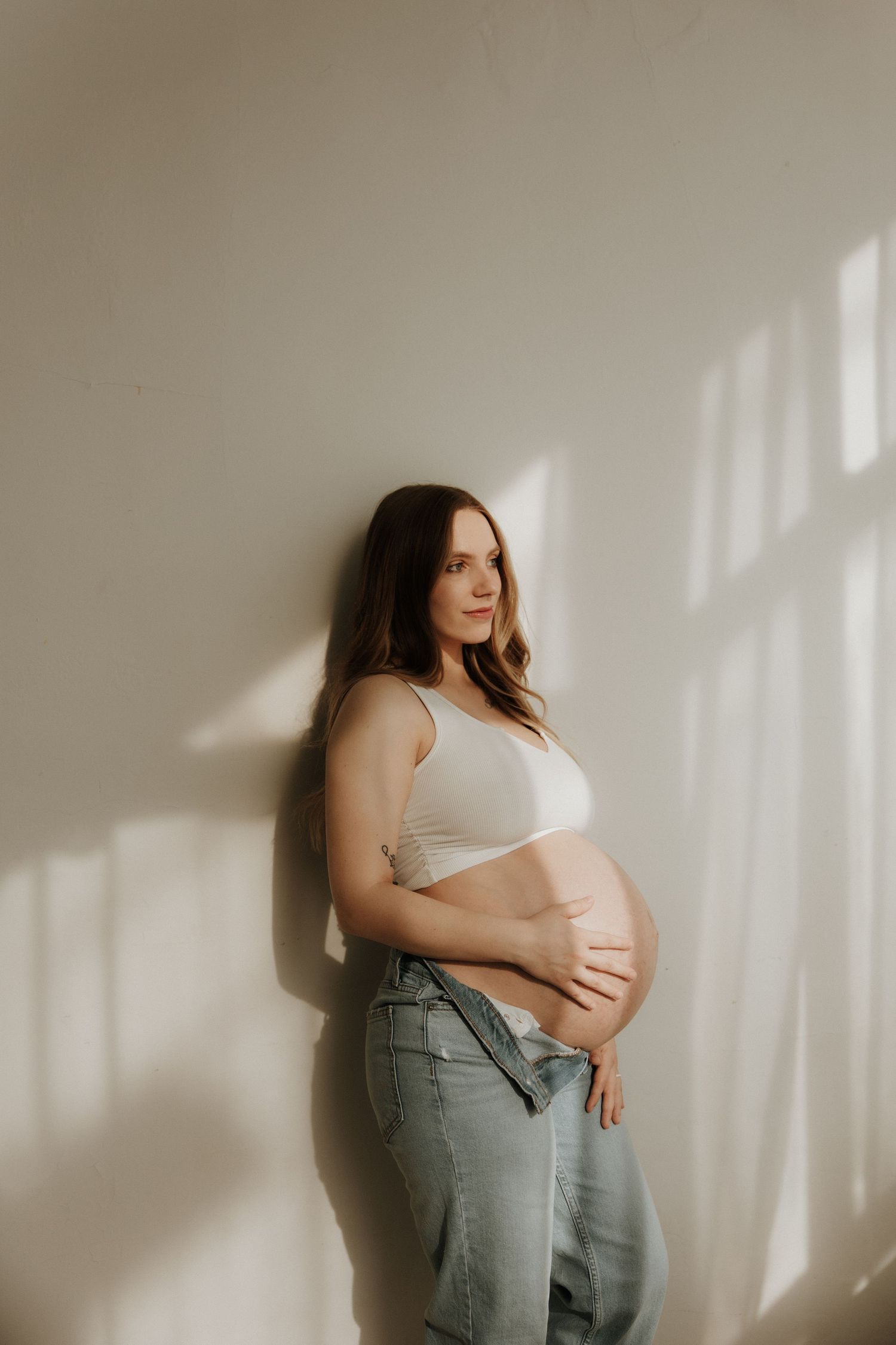 Minimalist maternity photos showing a pregnant figure in white bra and jeans against a light wall with window shadows.