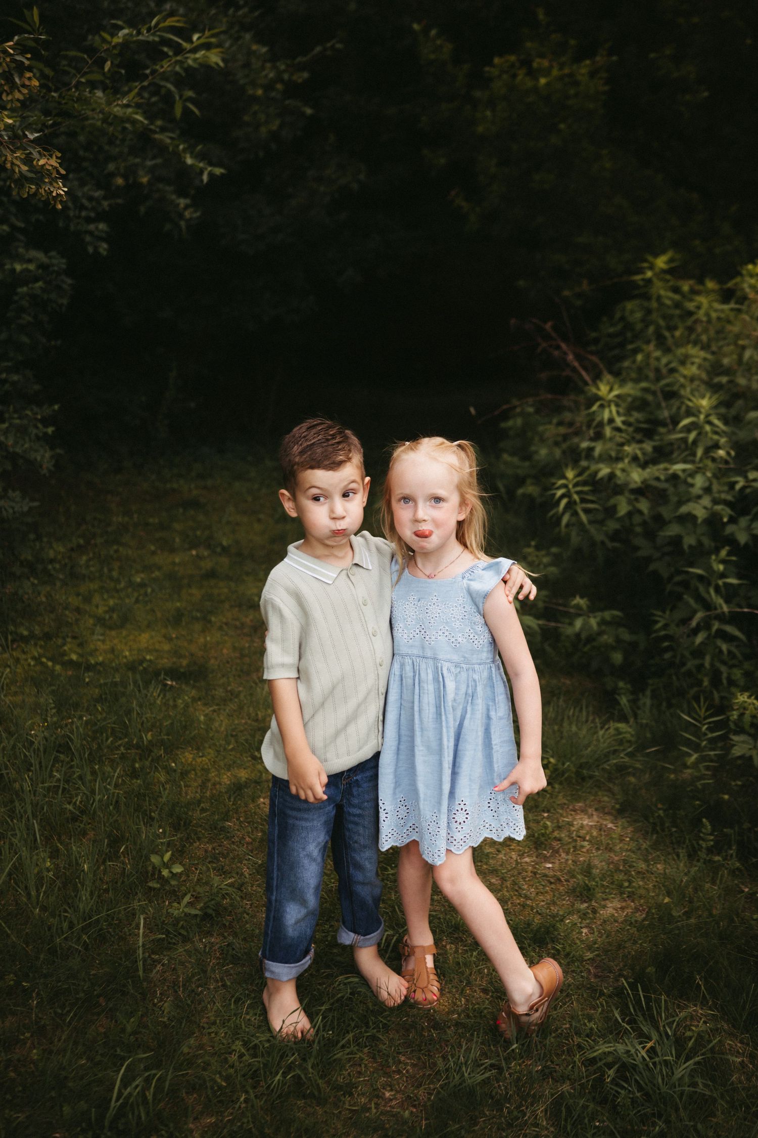 Two young children stand barefoot together in a wooded area wearing casual summer clothing.