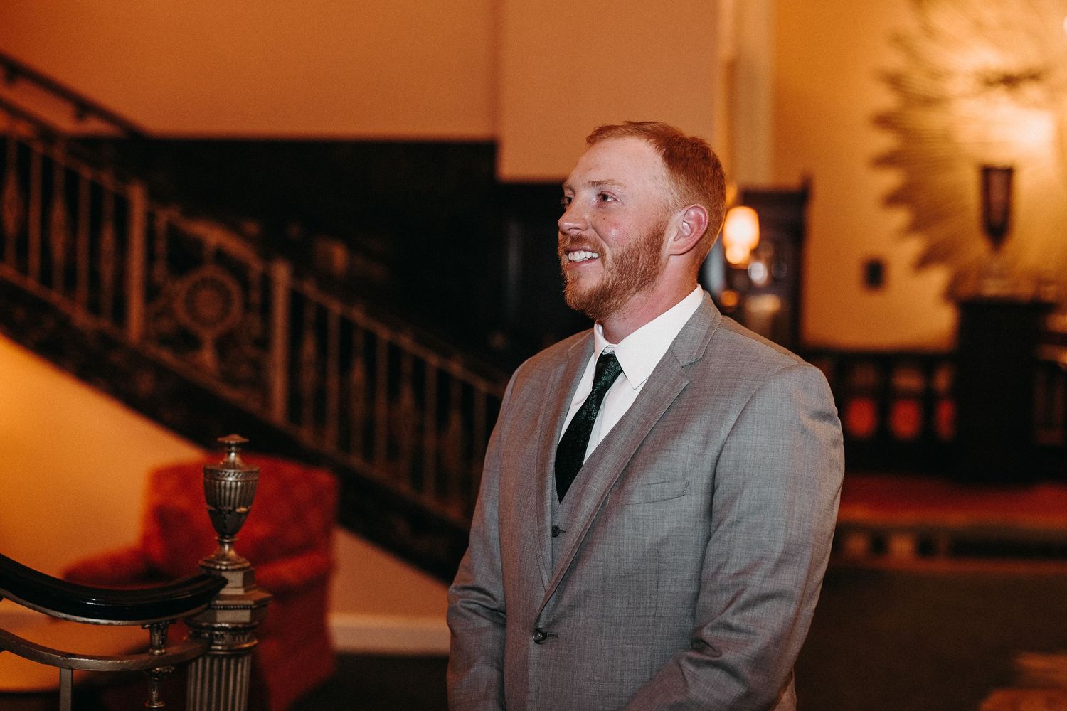 Person in grey suit smiling in an elegant venue with ornate staircase and warm lighting.