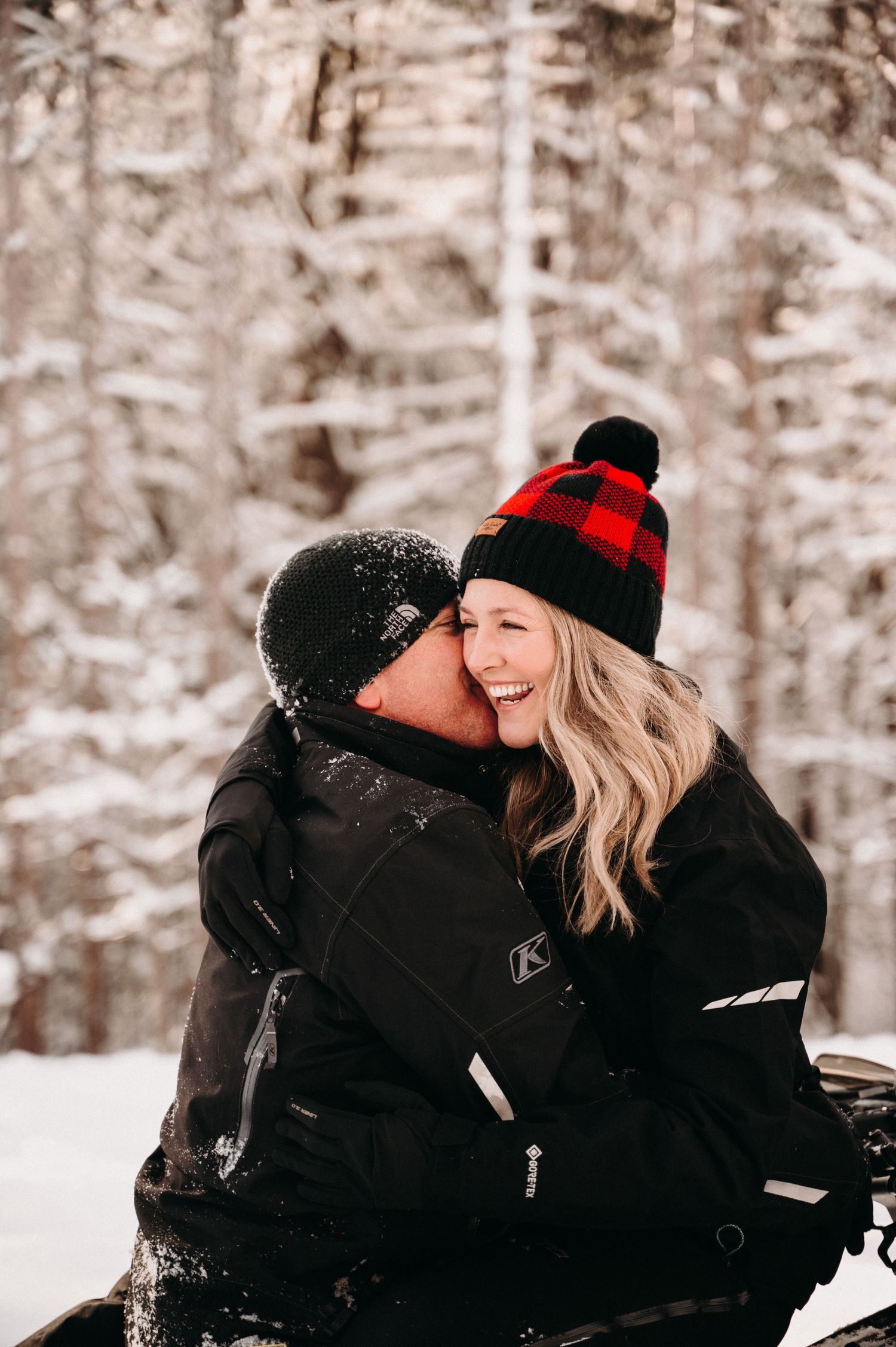 Couple sharing laughter while wearing winter coats and hats in snowy woods.