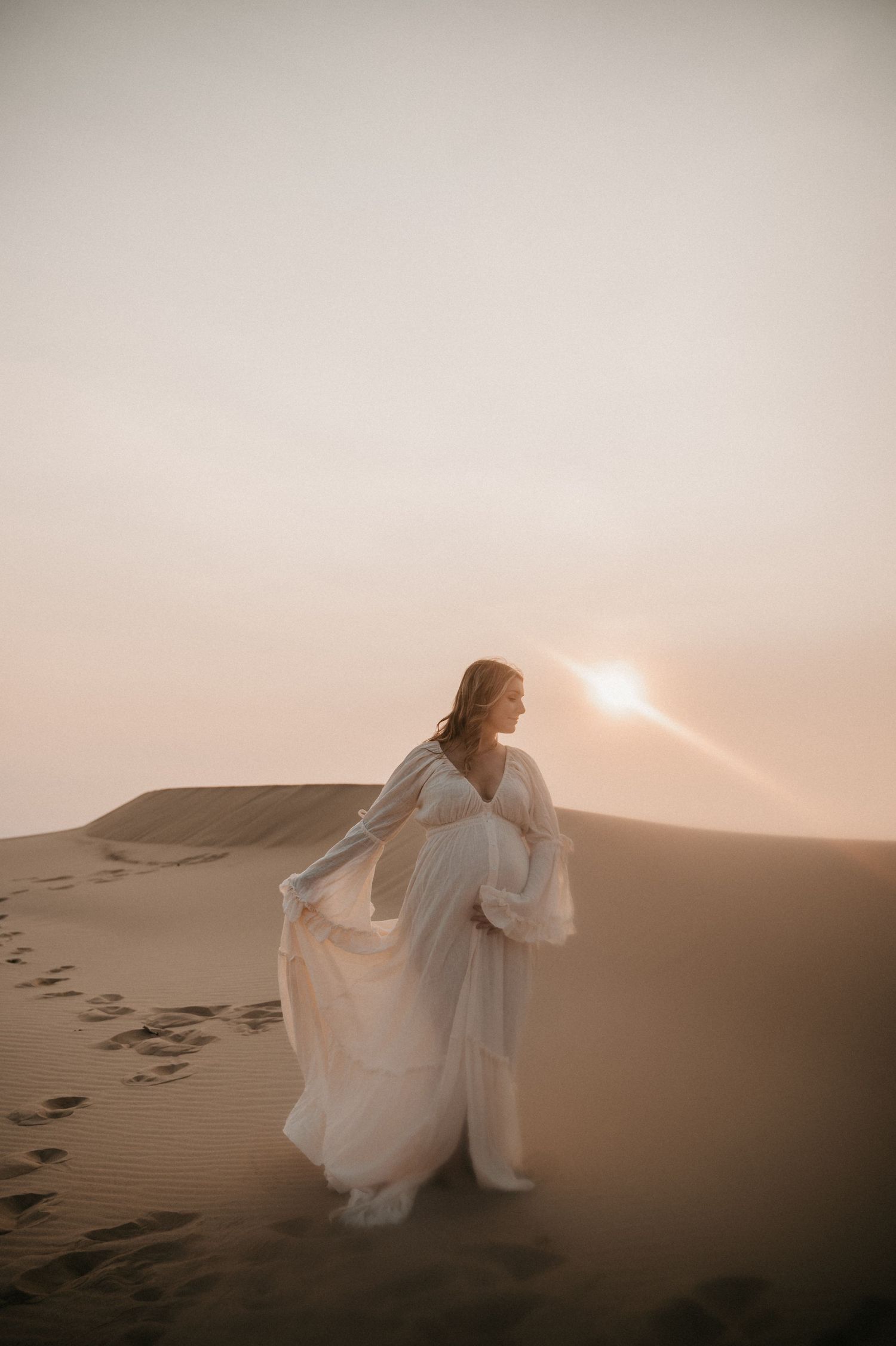 A flowing white dress billows in the desert wind as a figure stands against the setting sun on sandy dunes.