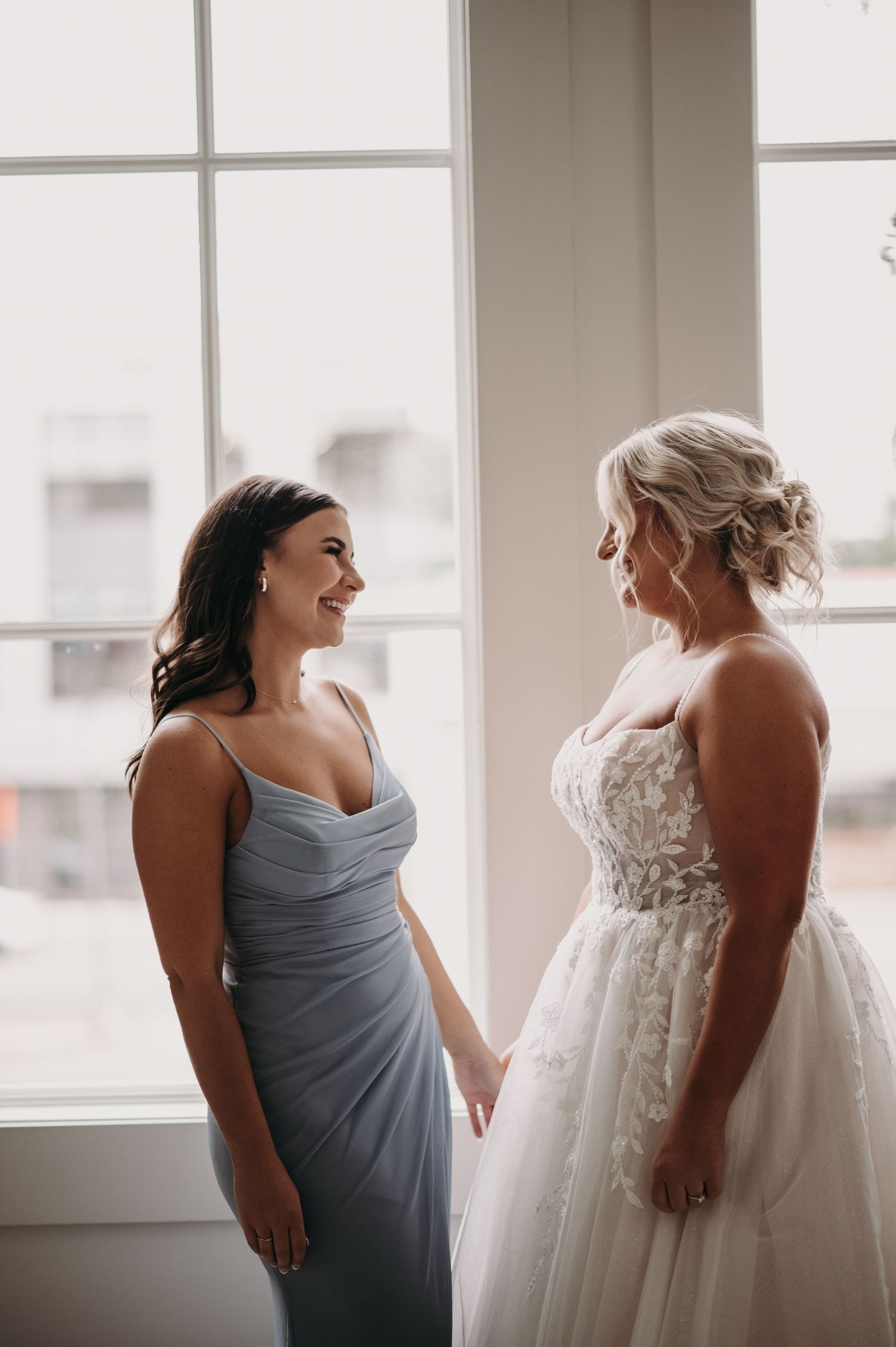 A bride in a white lace gown stands with a bridesmaid in a dusty blue dress by a bright window.