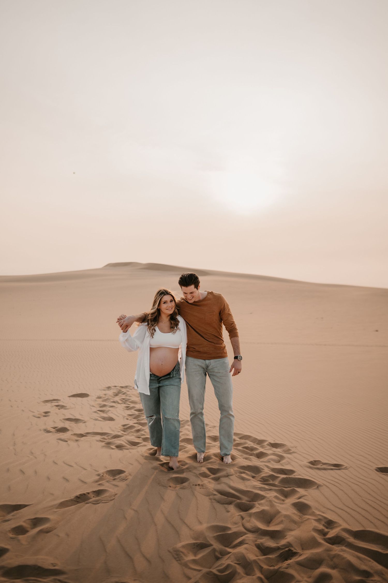 Couple walking hand in hand across desert sand dunes during sunset.