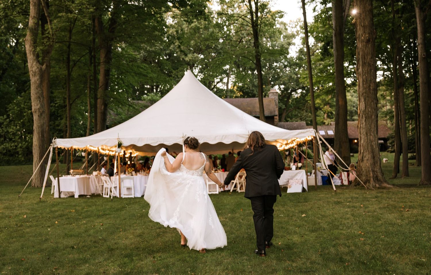 An outdoor wedding reception setup with white tents and tables arranged in a wooded area.
