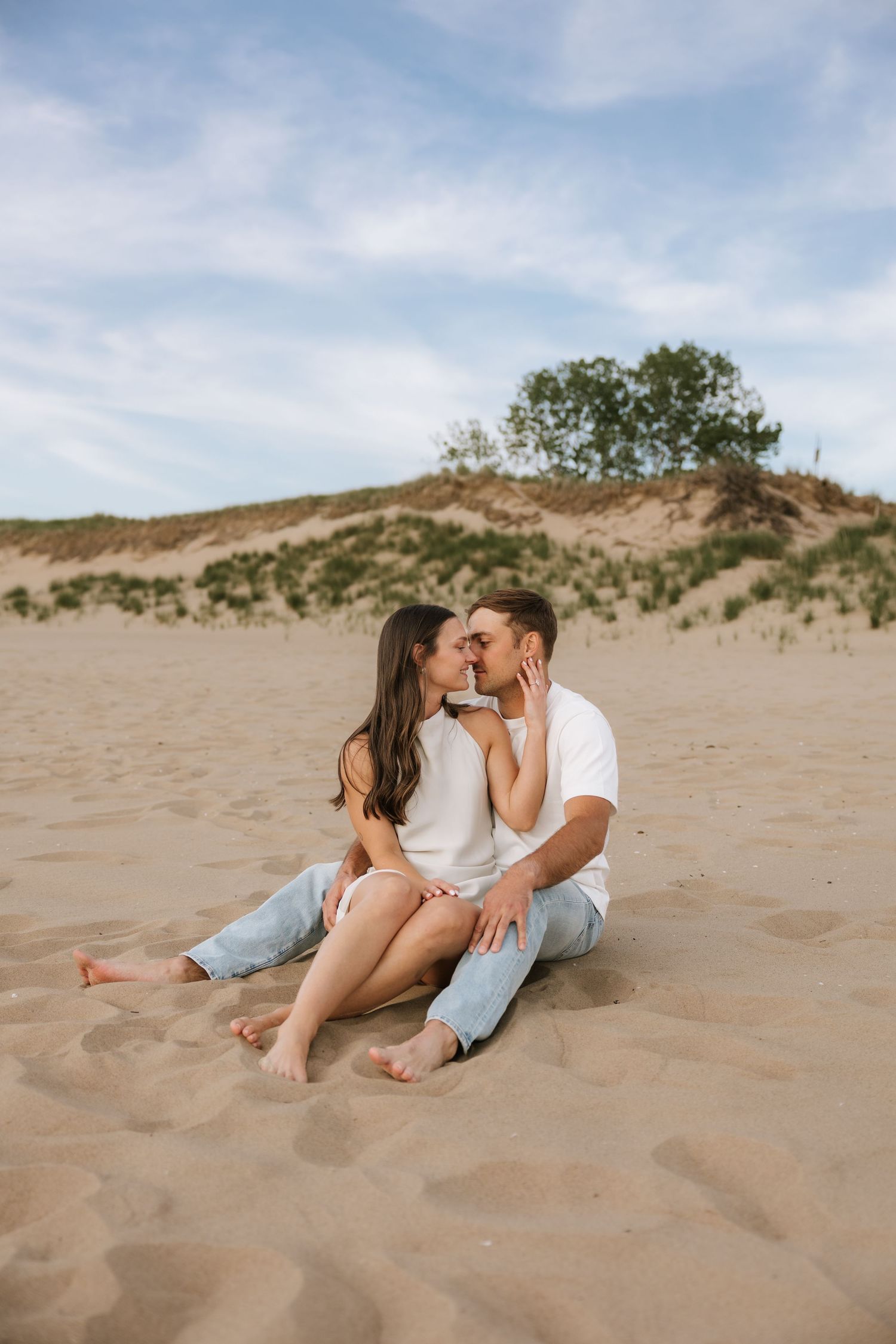 Couple sharing a kiss while sitting barefoot together on sandy beach dunes.