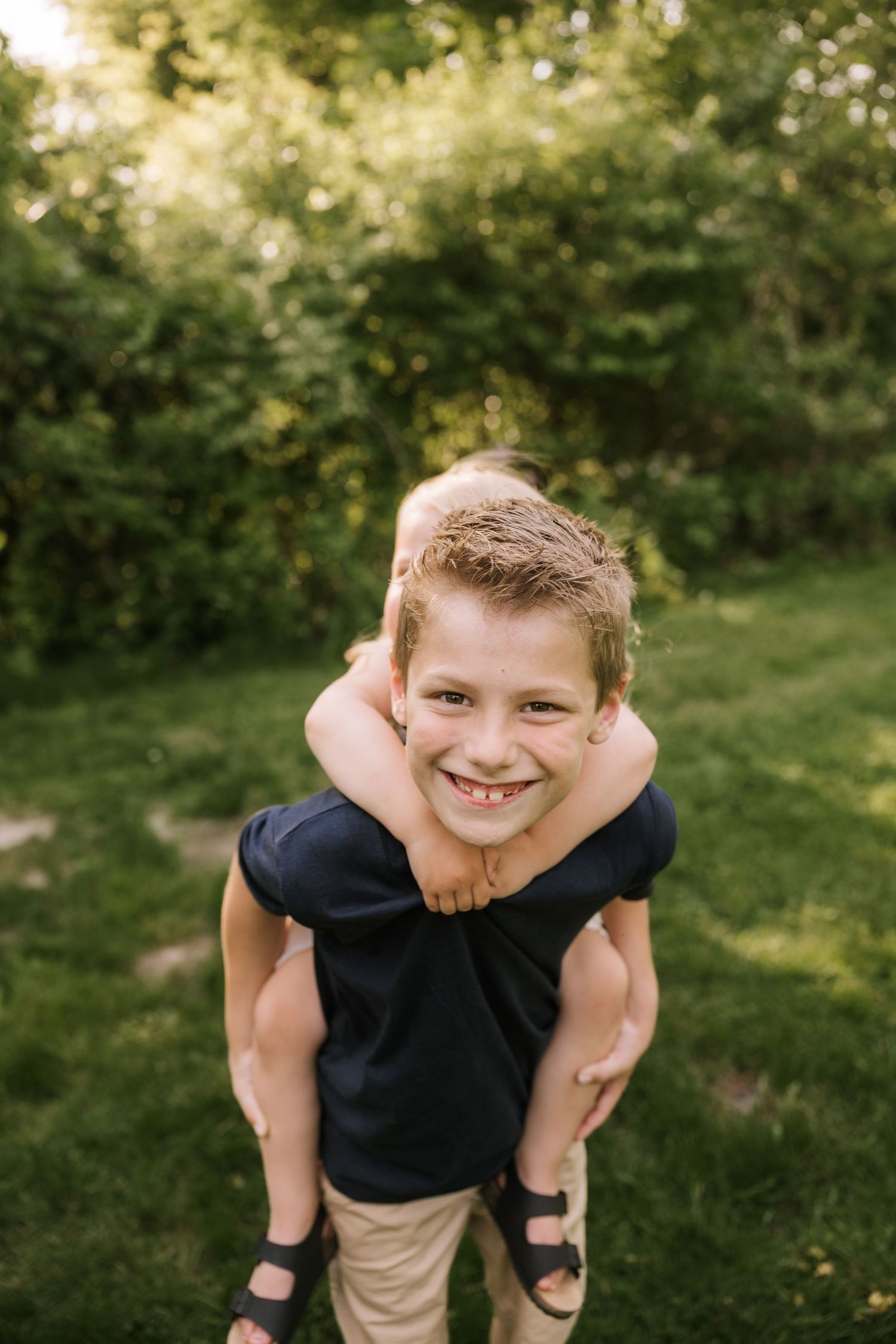 Smiling boy giving piggyback ride outdoors on green grass, wearing navy shirt and sandals.