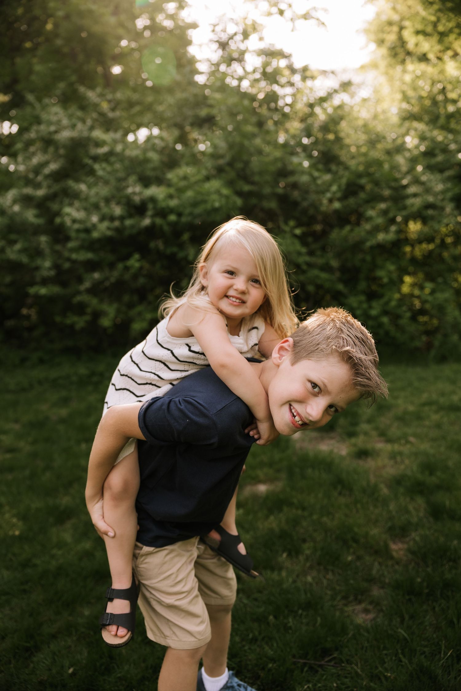 Young boy giving his little sister a piggyback ride outdoors in a sunny green garden.