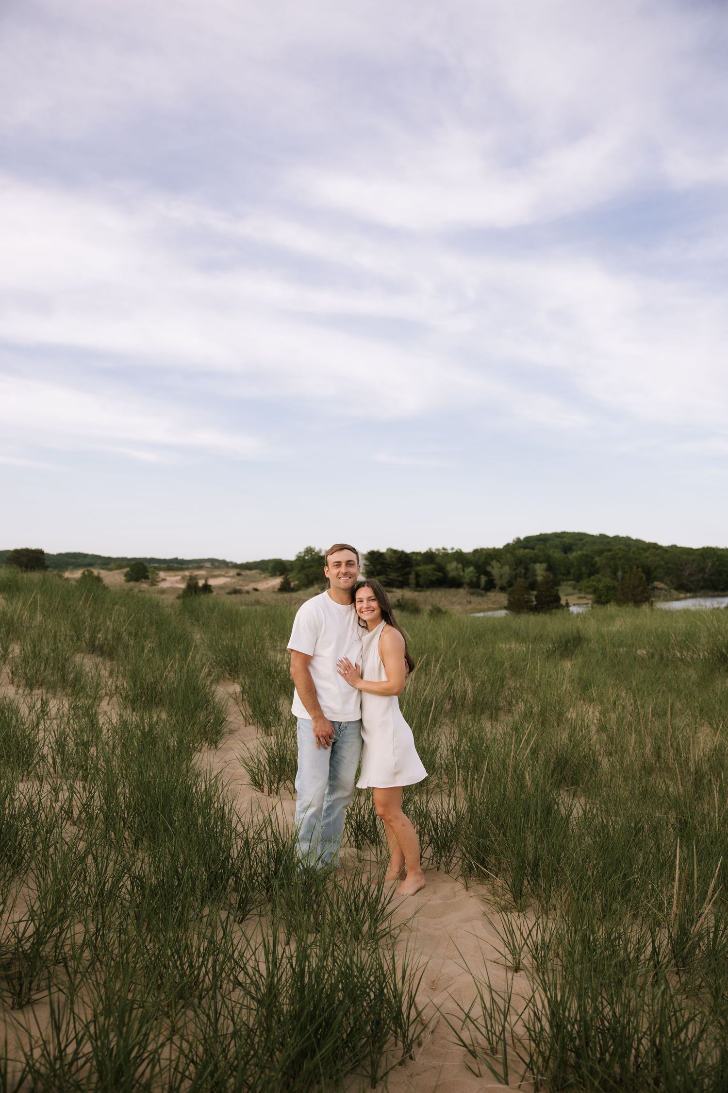 Happy couple embracing in sandy coastal dunes surrounded by tall green grass under cloudy sky.