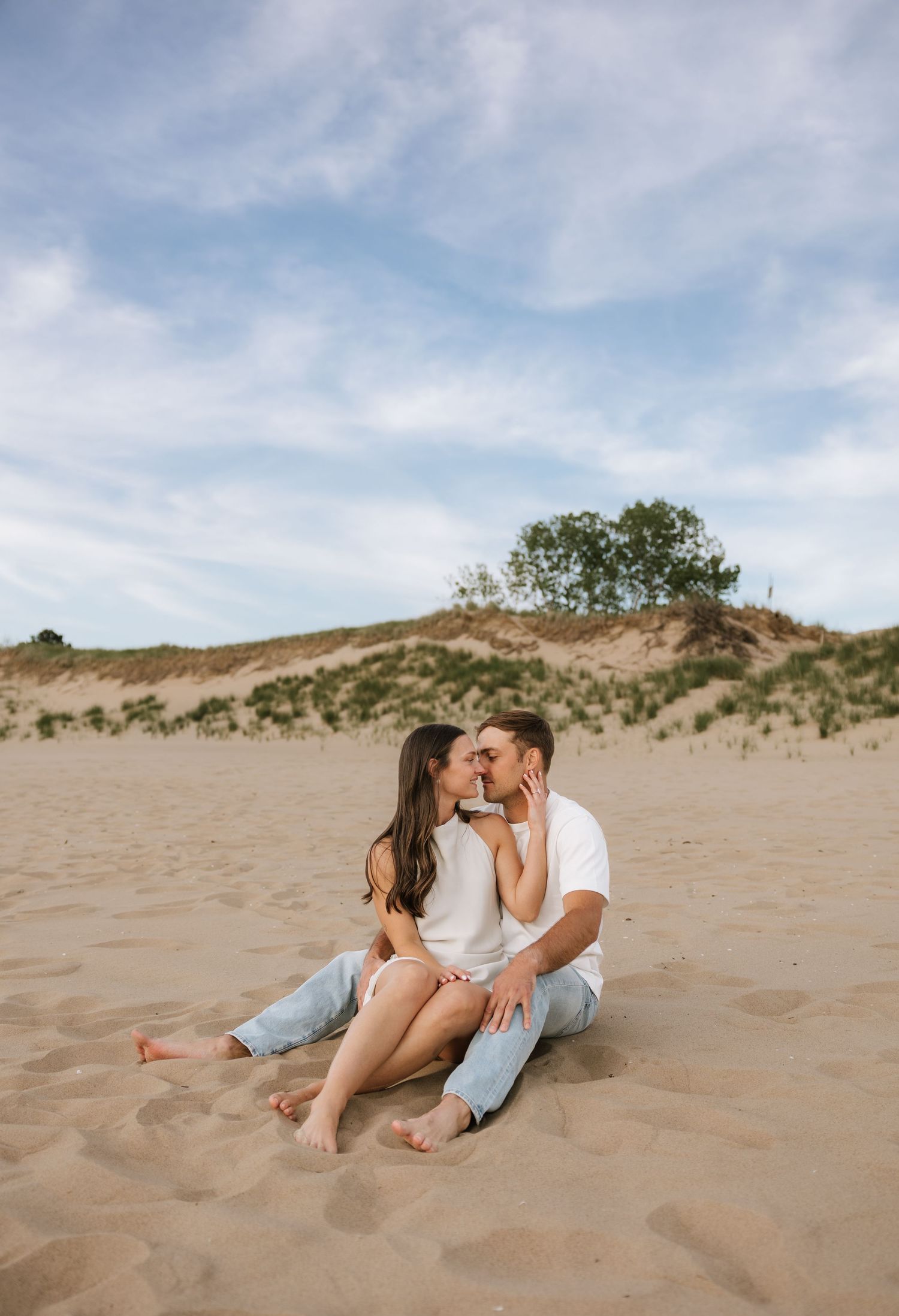 Couple kissing while sitting on sandy beach dunes under a blue sky during engagement photo session.