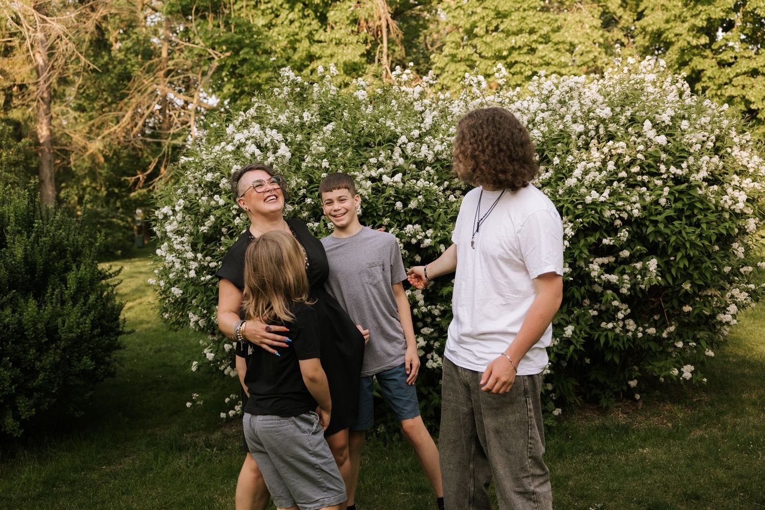 Group of four teenagers laughing and socializing outdoors near white flowering bushes on a sunny day.