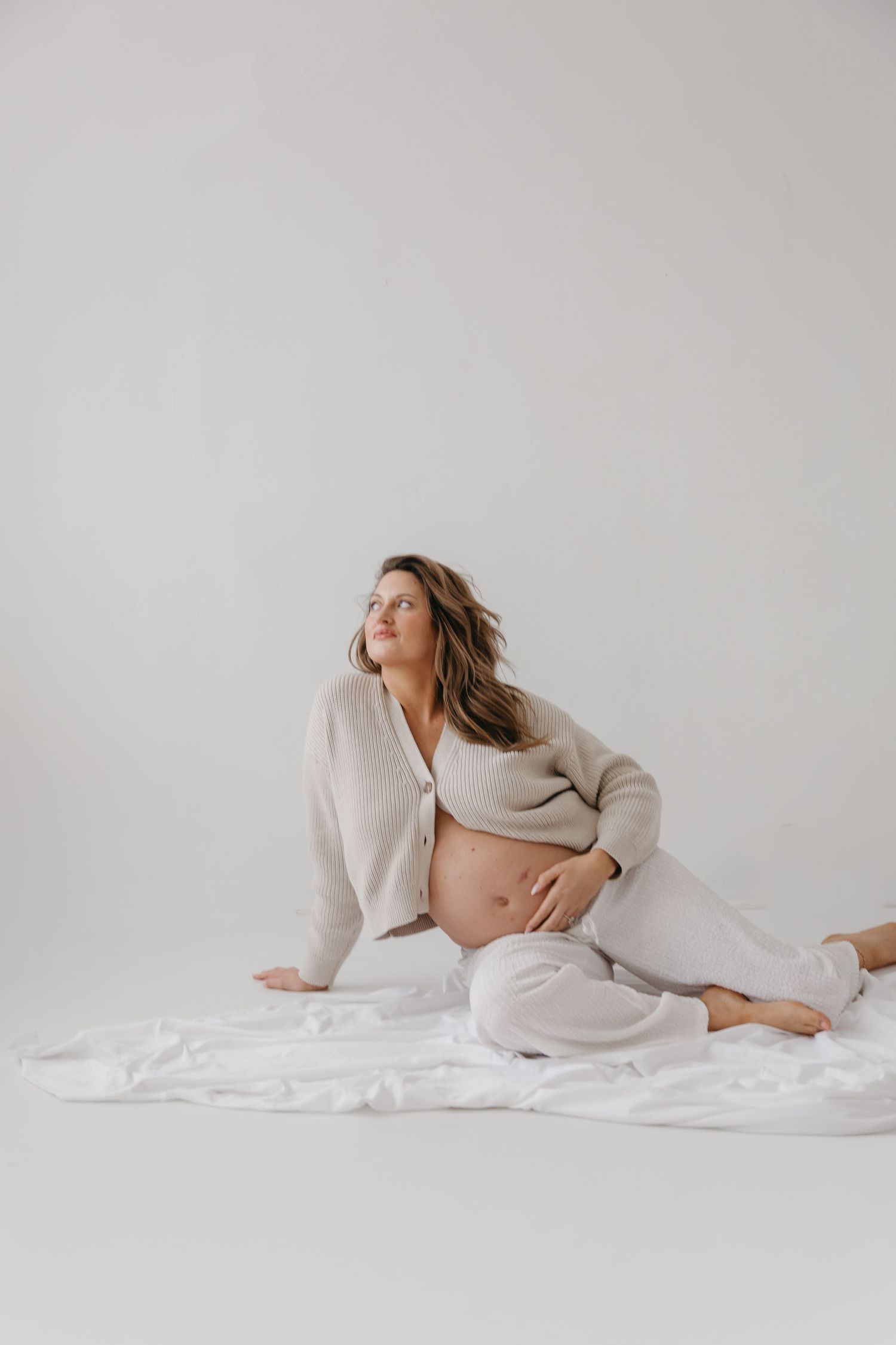 Pregnant woman in beige cardigan and white pants sitting on white fabric, cradling her baby bump.