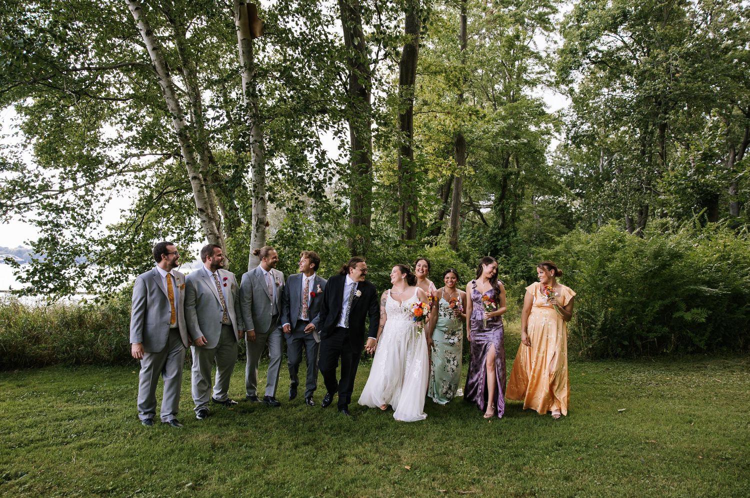 Wedding party and guests gathered for group photos on a grassy lawn with trees in background.