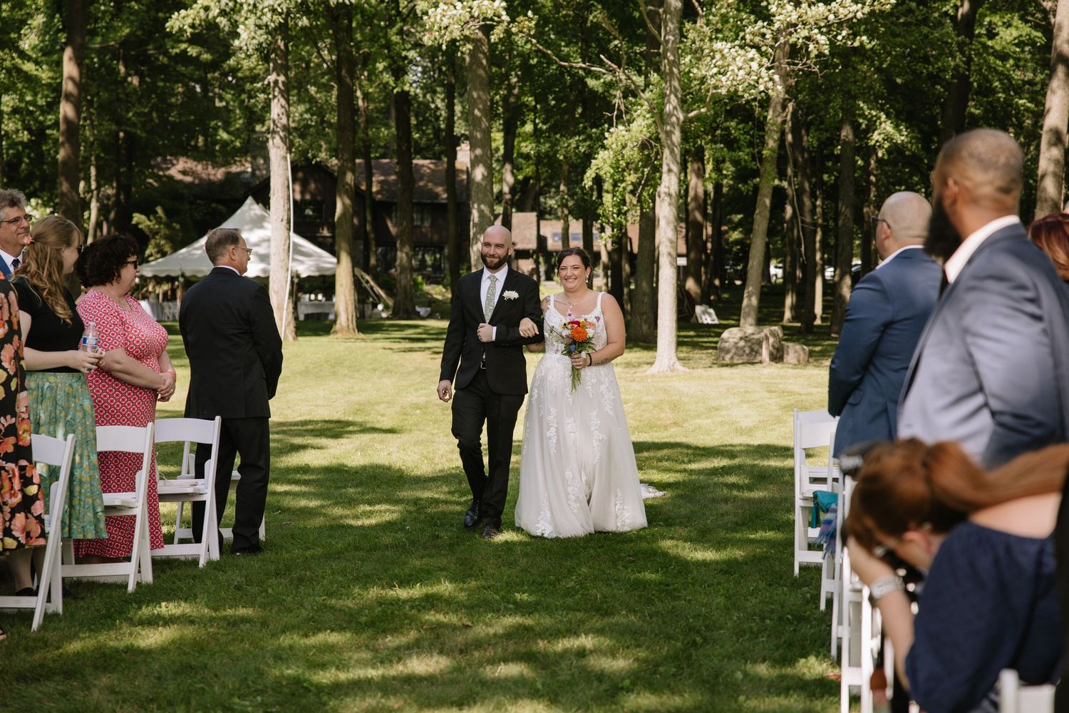 Guests gather outdoors at a sunny garden wedding ceremony with white chairs arranged on a green lawn.